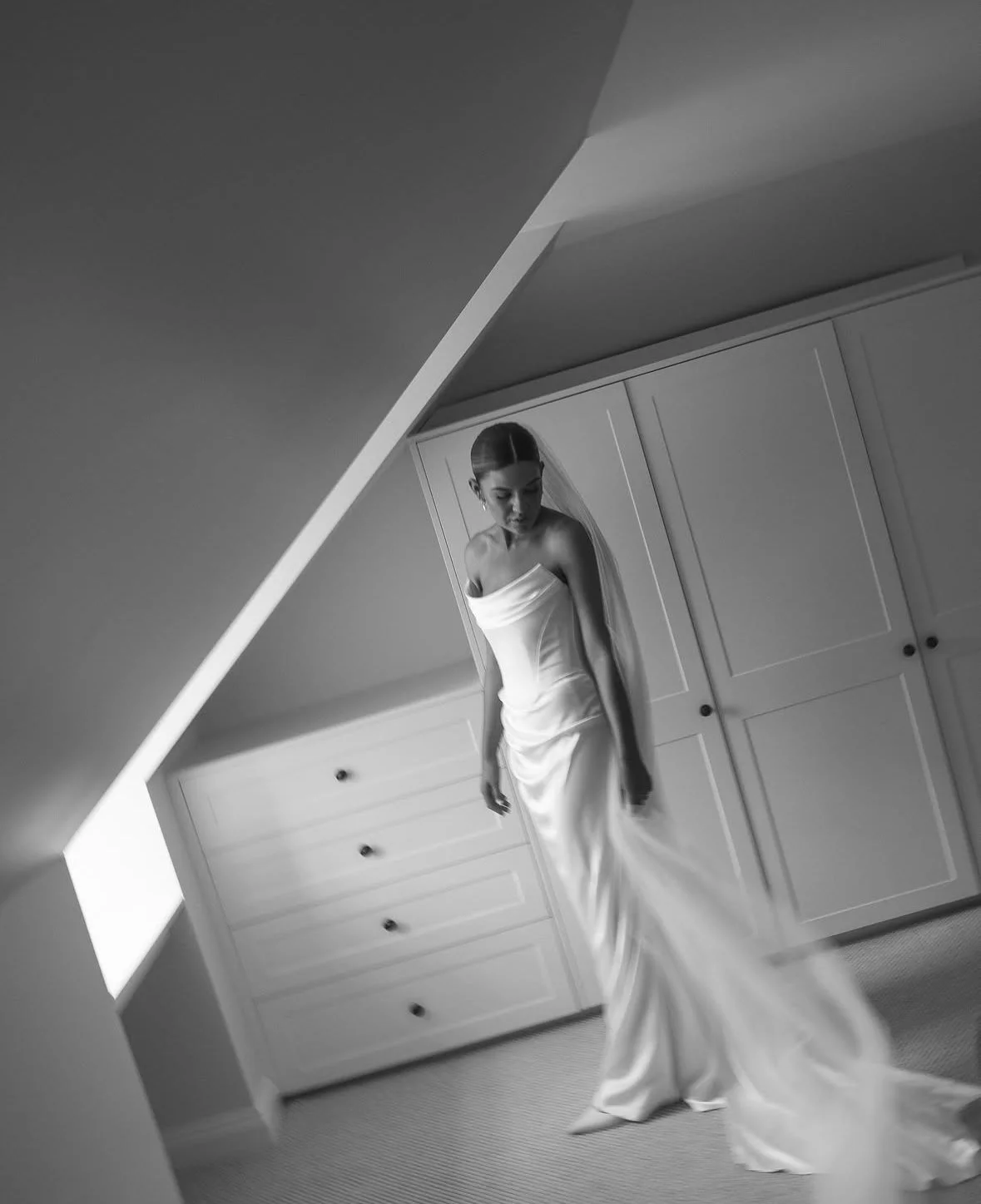 A woman in a satin wedding dress standing indoors near a white wardrobe, captured through a slanted mirror.