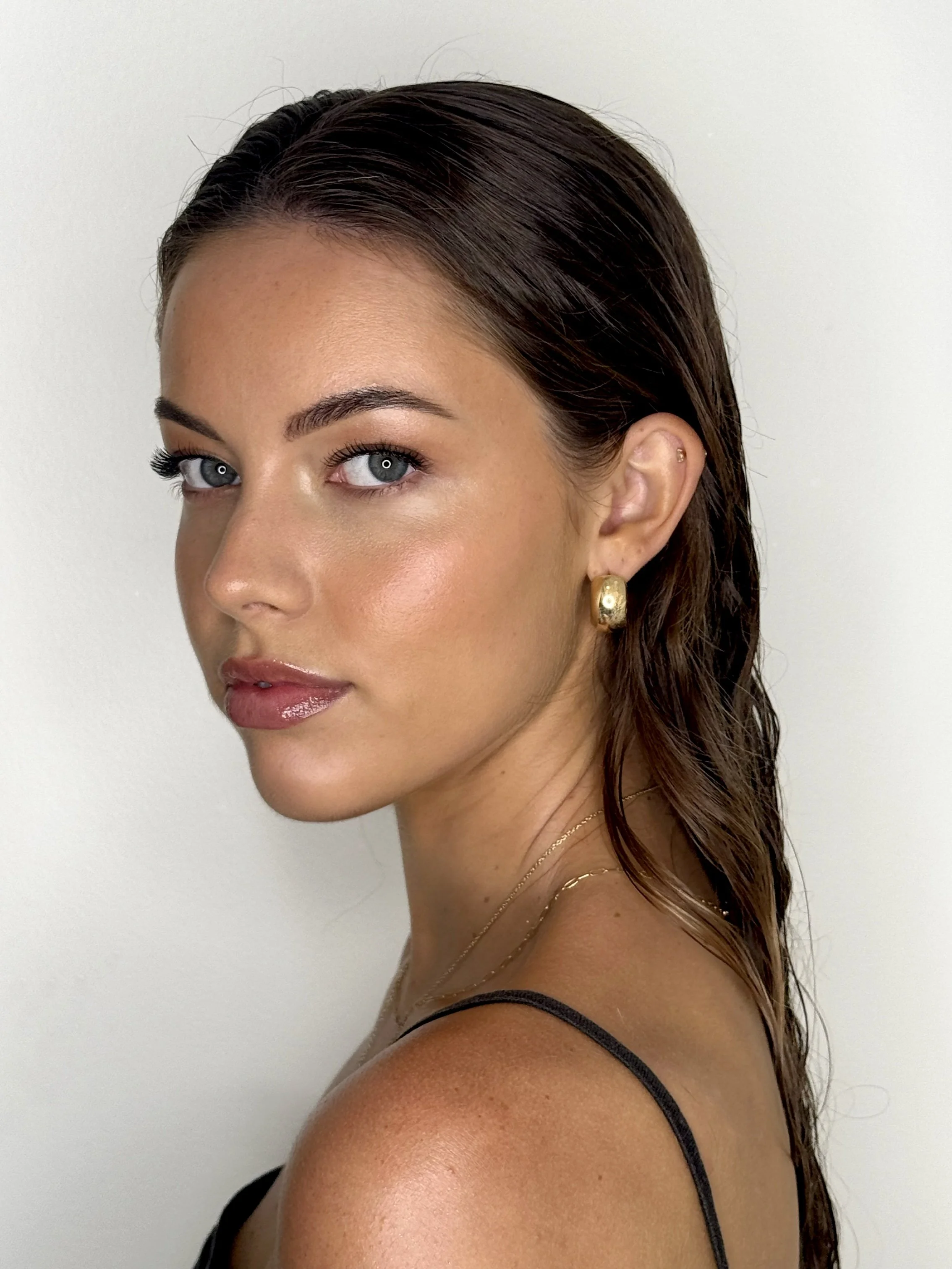 Close-up of a woman with wet brown hair, gold earring, and light makeup, looking over her shoulder with a neutral expression.