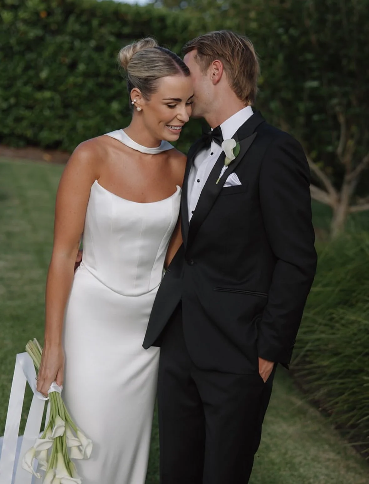 A bride and groom standing close together, smiling, outdoors with greenery in the background, the bride holding a bouquet of calla lilies.