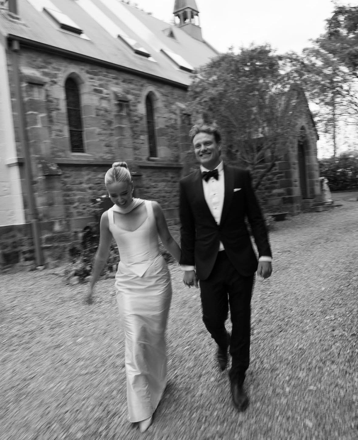 A smiling bride and groom walking hand in hand outside a stone church in black and white.