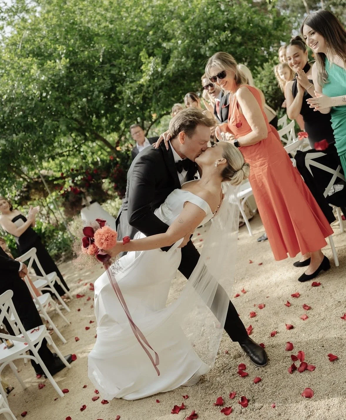 A newlywed couple sharing a kiss at their outdoor wedding ceremony surrounded by friends and family, with rose petals scattered on the ground.