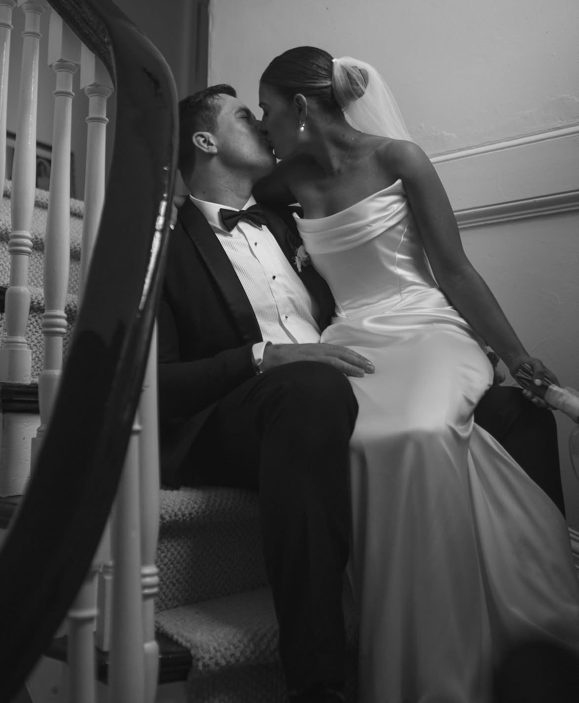 A bride and groom sharing a kiss on a staircase, with the bride sitting on the groom's lap. The scene is captured in black and white.