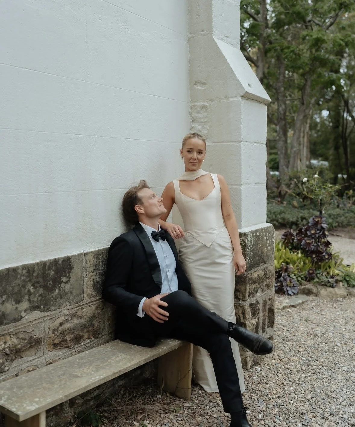 A man in a black tuxedo with a bowtie and a woman in a white evening gown are posing outdoors against a white and stone building wall. The man is sitting on a bench, looking up at the woman, who is standing beside him with her hand resting on his sho