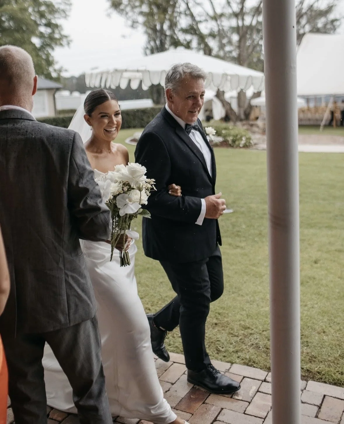 A bride in a white wedding dress holding a bouquet of flowers, smiling, as she walks with an older man in a black tuxedo holding her arm, during a wedding celebration outdoors on a cloudy day.