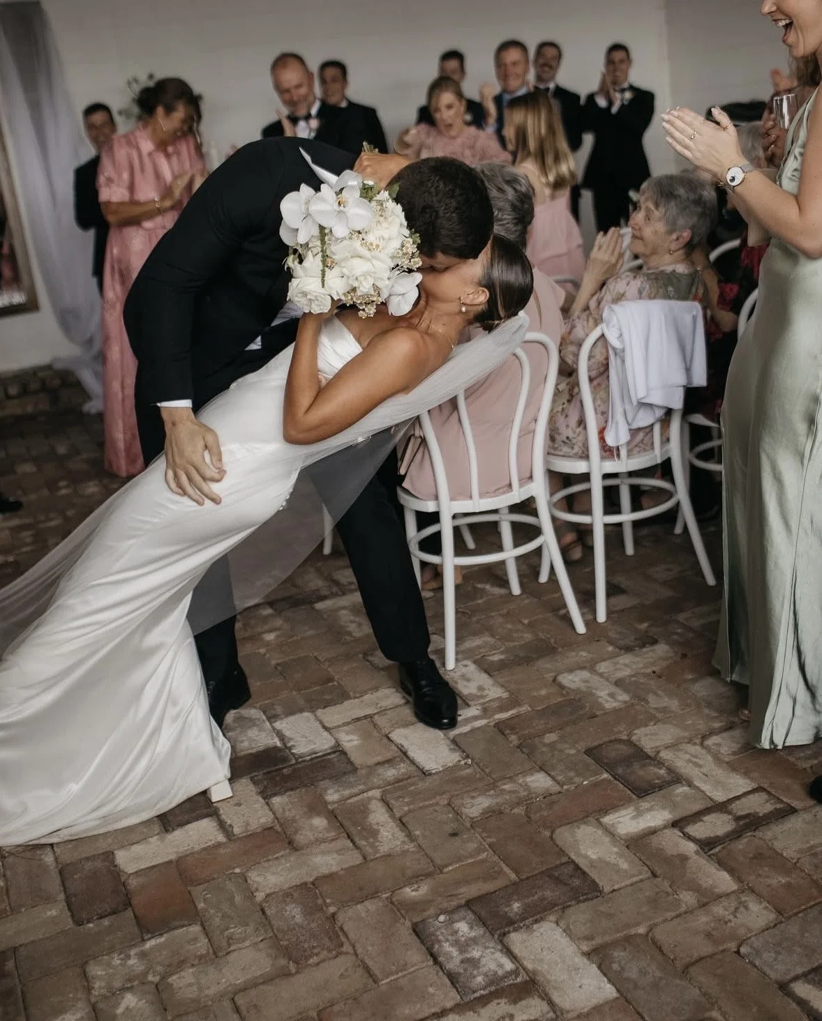 A newlywed couple sharing a kiss at their wedding reception, surrounded by guests applauding and celebrating.