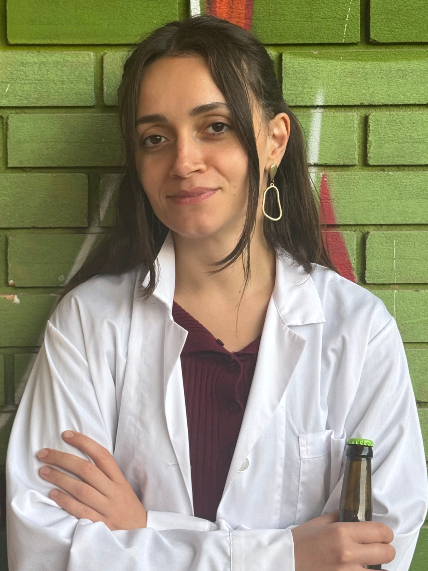 A woman with long dark hair, wearing a white lab coat and gold earrings, stands against a green brick wall with some graffiti. She is holding a brown glass bottle with a green cap in her right hand and has her arms crossed.