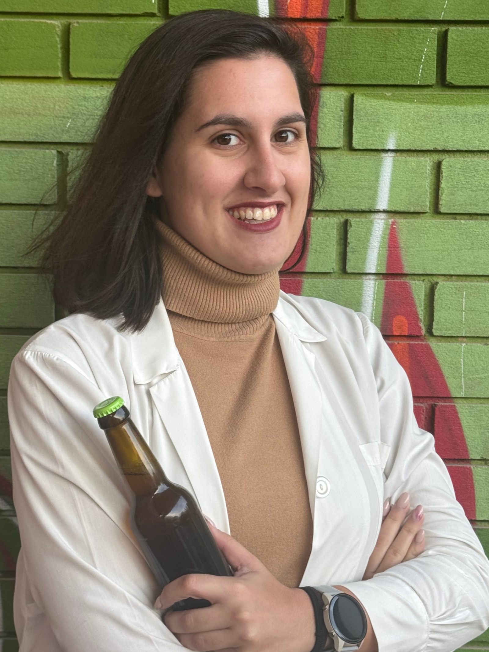 Smiling woman with shoulder-length dark hair wearing a beige turtleneck and white jacket, standing in front of a green brick wall with colorful graffiti.