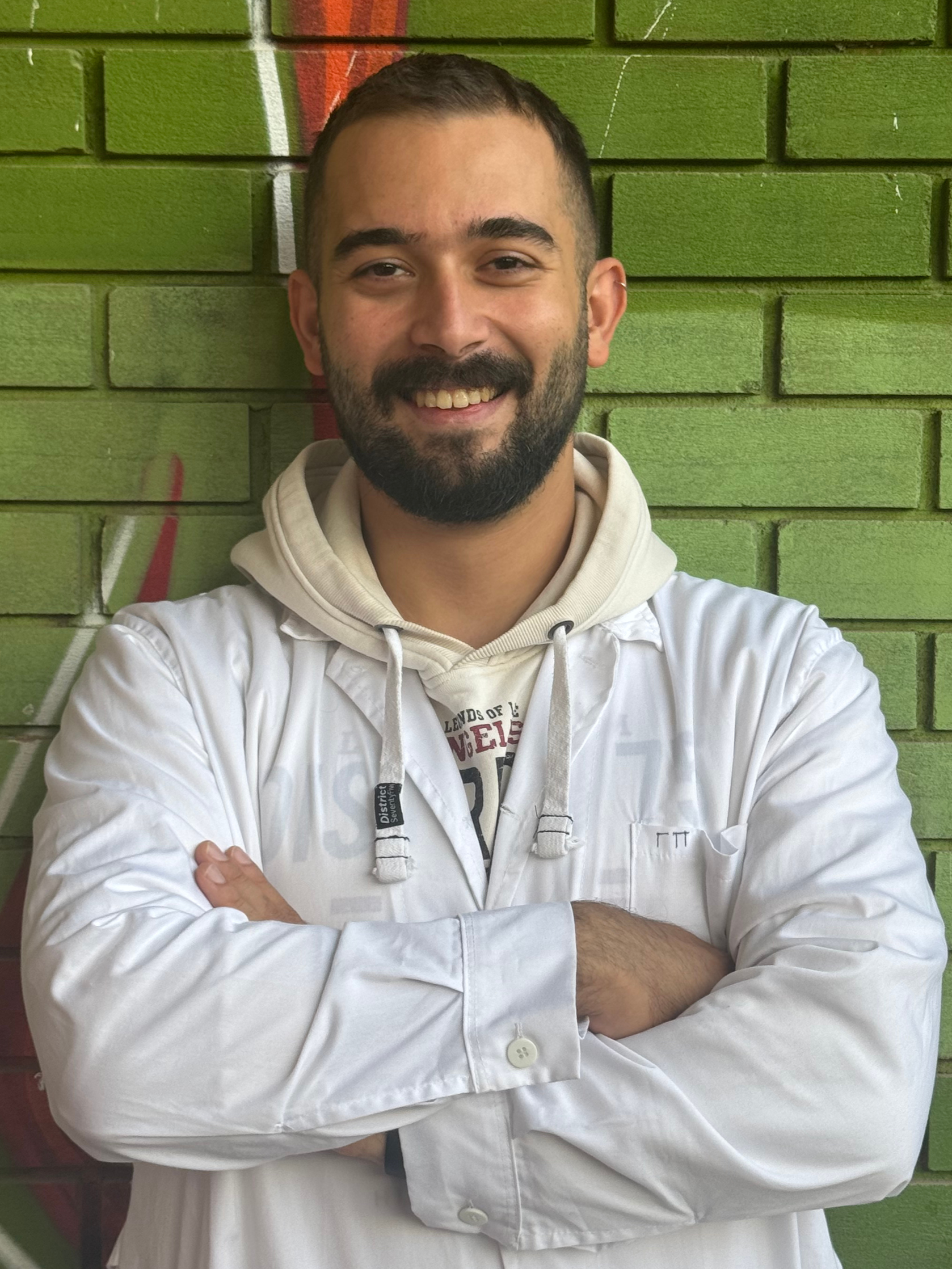 A smiling man with a beard and mustache, crossed arms, standing in front of a green brick wall with graffiti.
