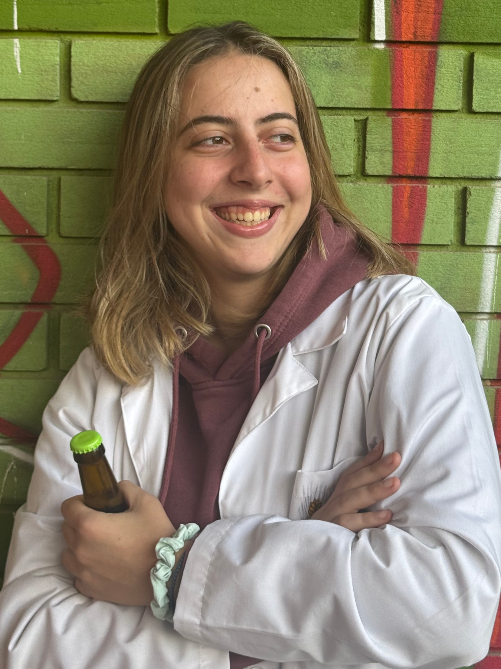 A young woman with shoulder-length light brown hair, smiling and crossing her arms while holding a brown glass bottle with a green cap. She is wearing a white lab coat over a maroon hoodie and standing against a green brick wall with red graffiti.
