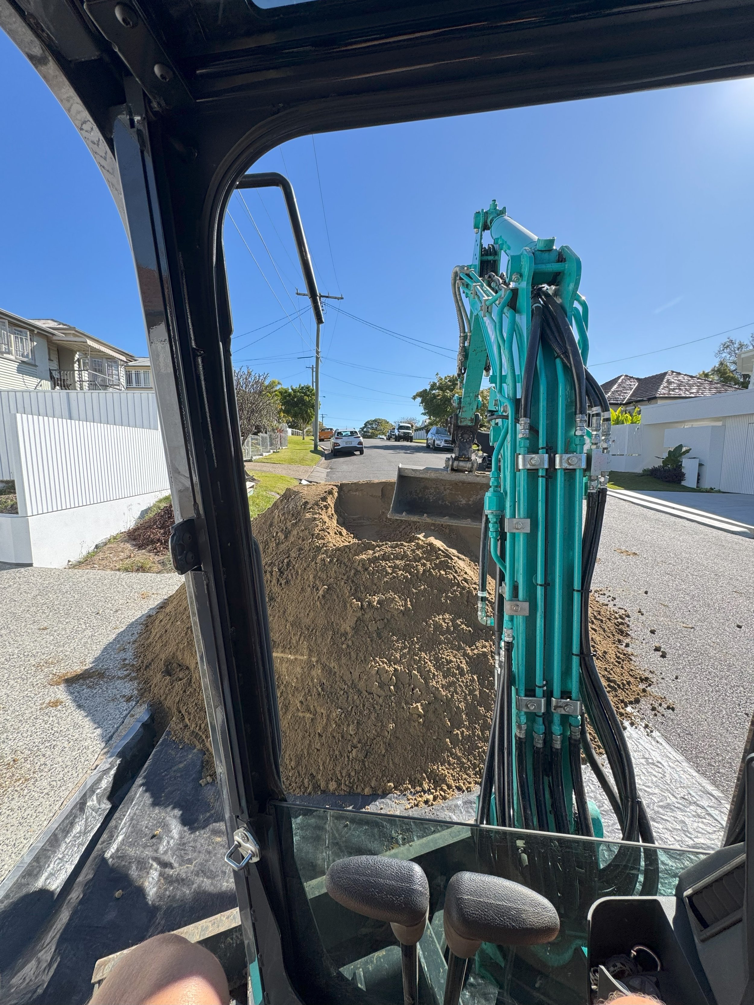 View from inside an excavator showing an excavator bucket filled with dirt on a residential street with parked cars, houses, and utility poles.