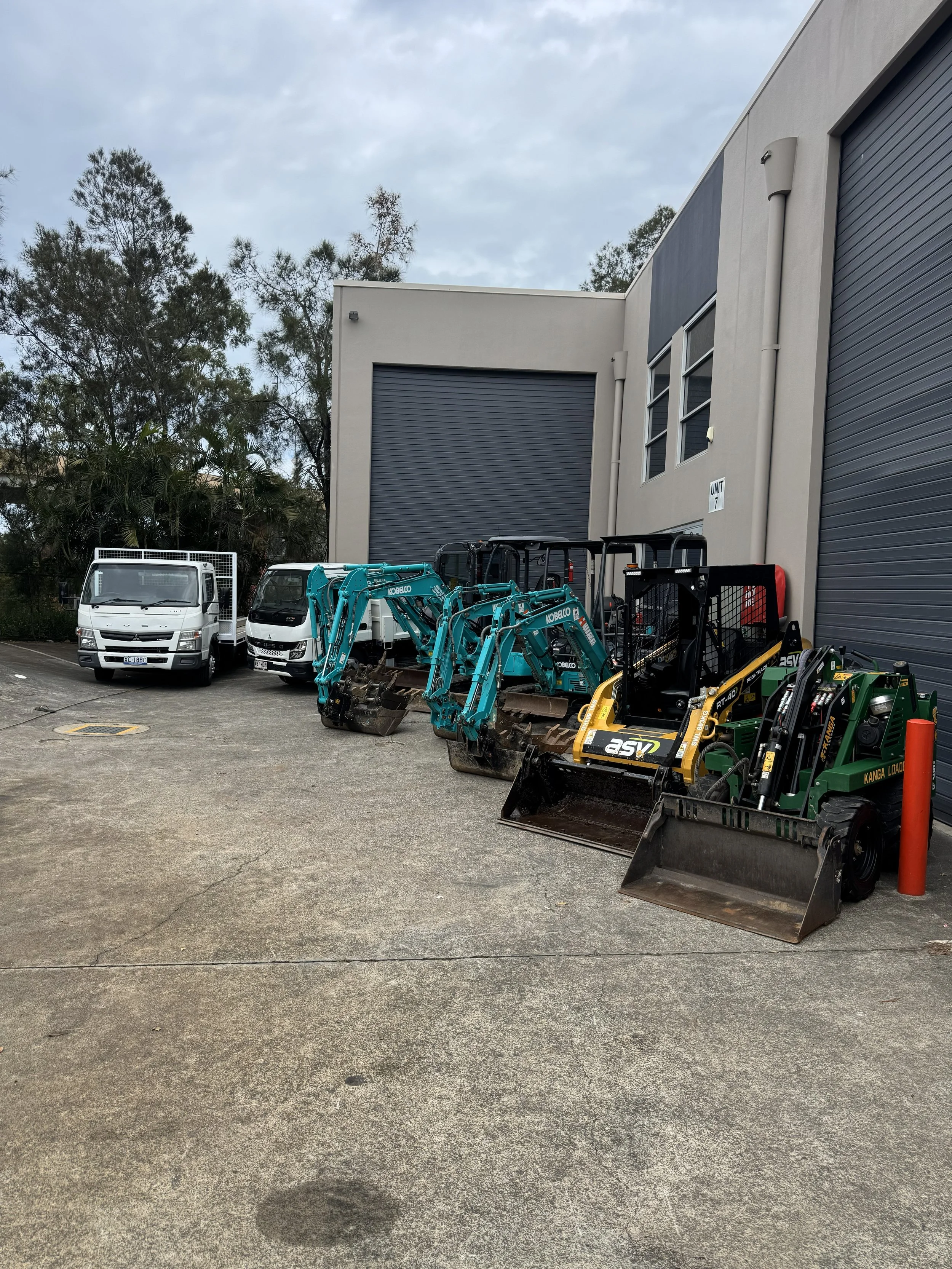 A parking lot with three white trucks and multiple small construction vehicles, including mini excavators and skid-steer loaders, parked outside a modern industrial building with large gray roller doors.