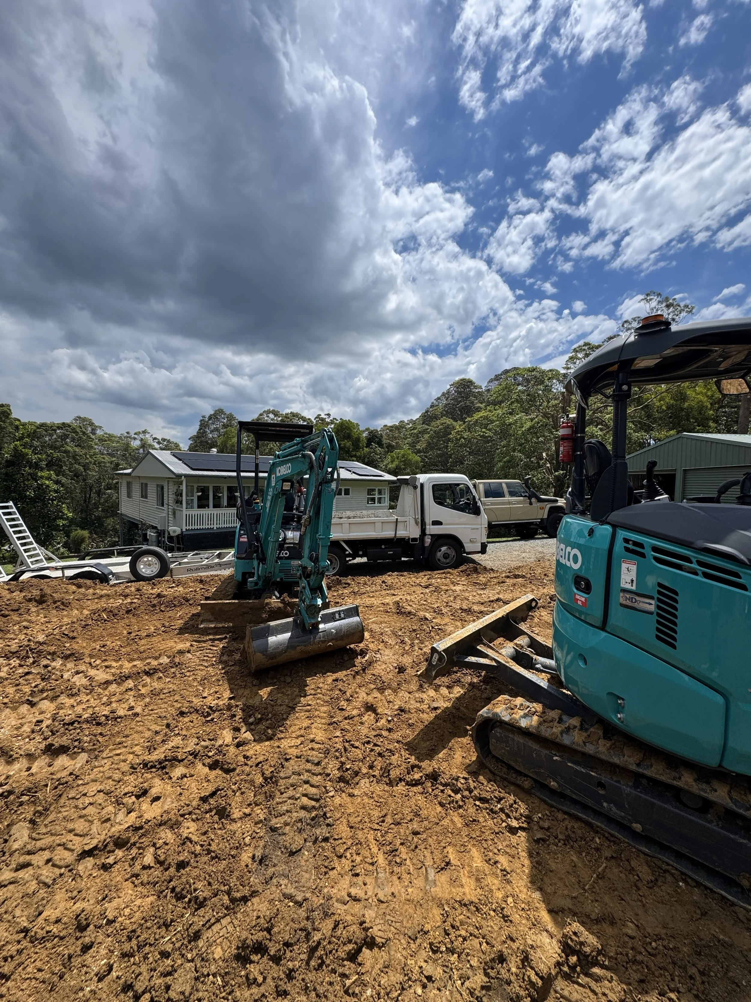 Construction site with a small excavator and a mini bulldozer on a dirt ground, residential houses and trees in the background, partly cloudy sky.