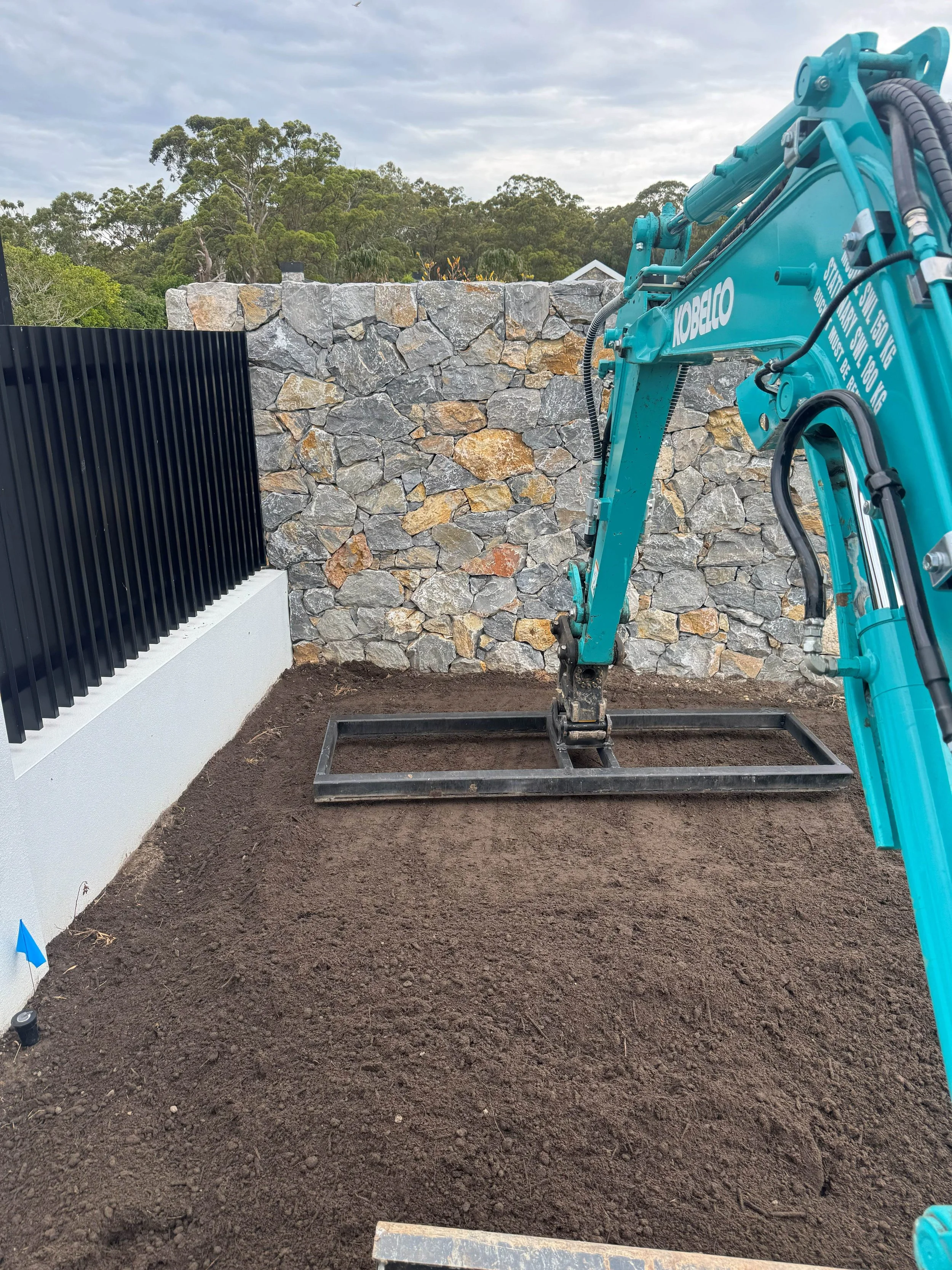 Construction site with a small blue excavator digging into the dirt near a stone wall and black fencing.