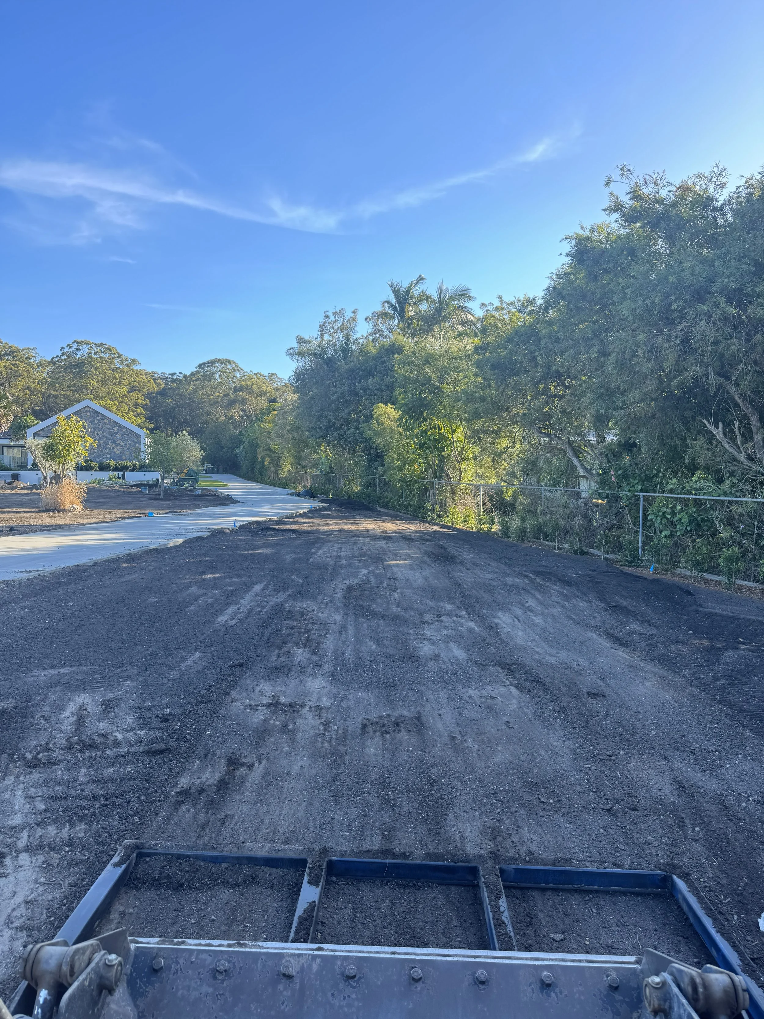 View from a construction vehicle on a partially paved road with trees and a house in the background under a partly cloudy blue sky.