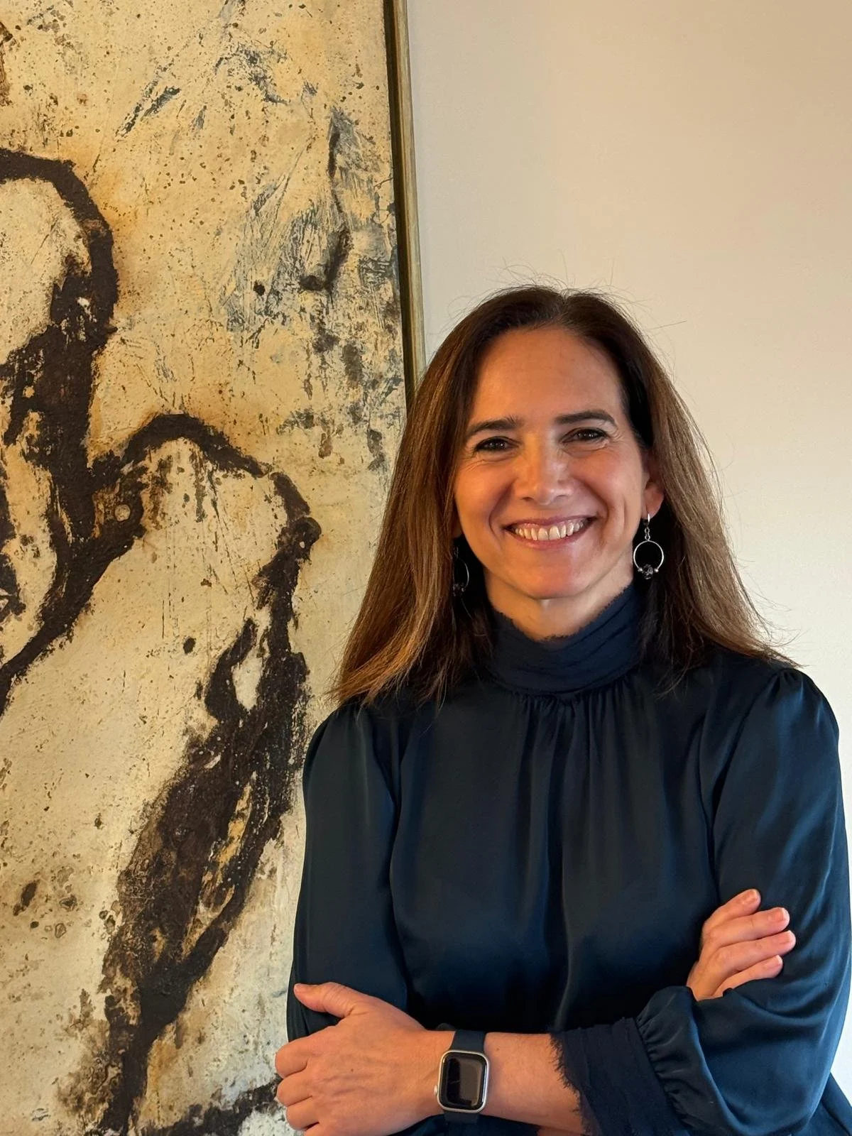 Mujer sonriendo con brazos cruzados en interior, lleva reloj inteligente y aretes, fondo con cuadro abstracto.