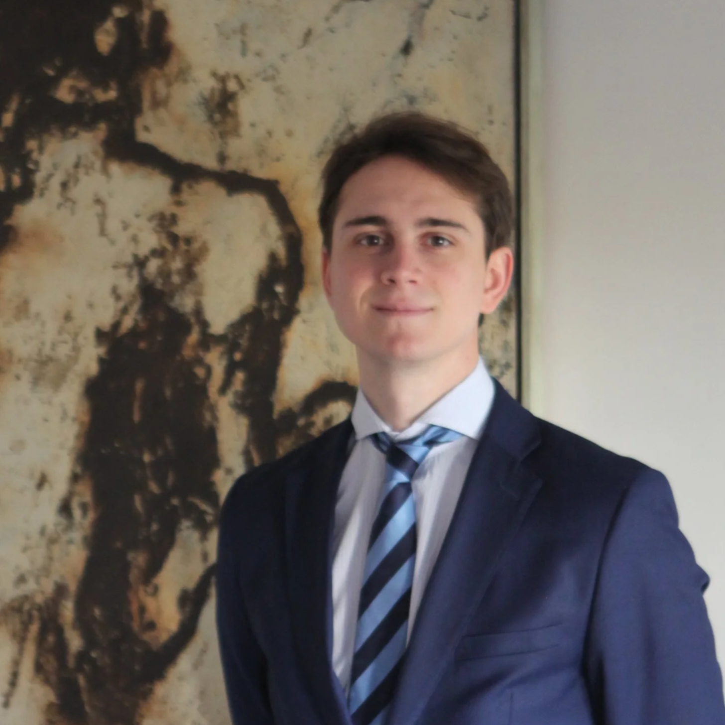 Joven vestido con traje oscuro y corbata a rayas, posando en un lugar con arte en la pared de fondo.