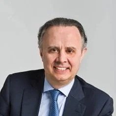 Hombre con cabello peinado y corbata, vestido con saco y camisa formal, sonriendo frente a fondo gris.