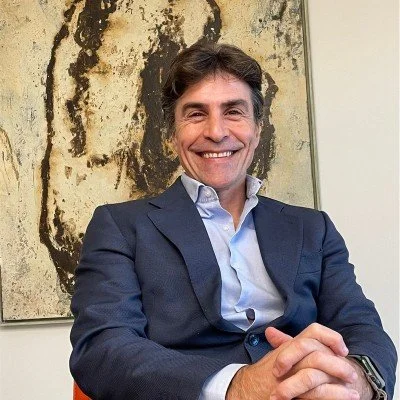 Hombre sonriente con traje oscuro y camisa blanca, sentado en una oficina, en un fondo con una pintura abstracta.