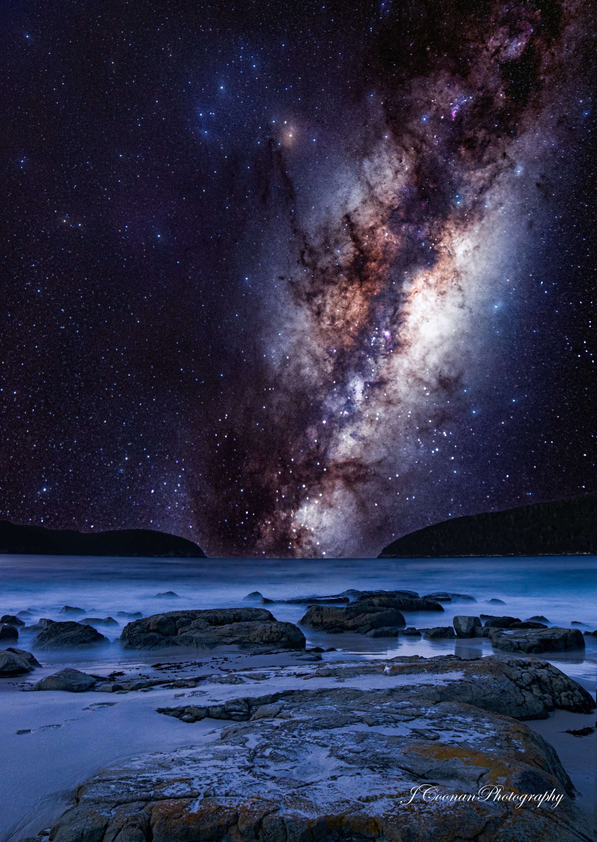 Night sky over ocean with visible Milky Way galaxy and stars, rocky shoreline in foreground.