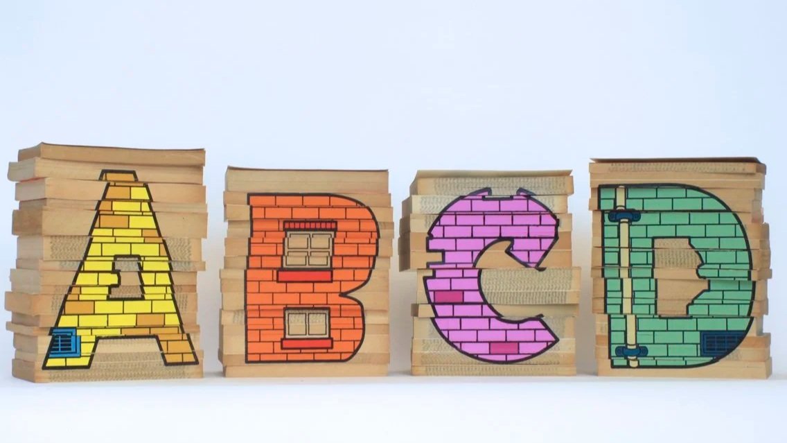 Stacked wooden blocks spelling out the letters 'A', 'B', 'C', 'D' with painted brick patterns in yellow, orange, purple, and green.