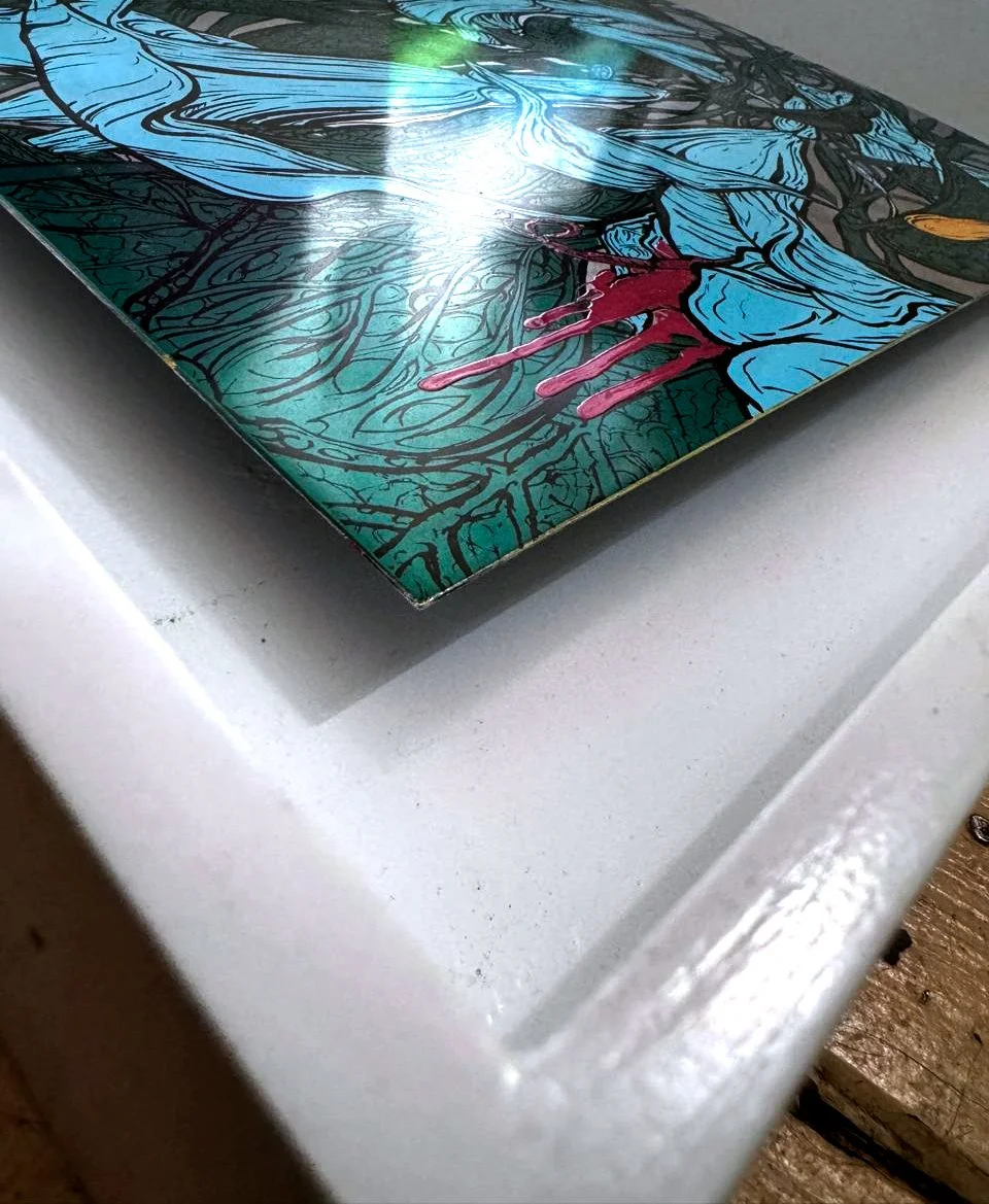 Close-up of a colorful, abstract artwork on a metallic surface with a white border, featuring blue, green, red, and black lines.