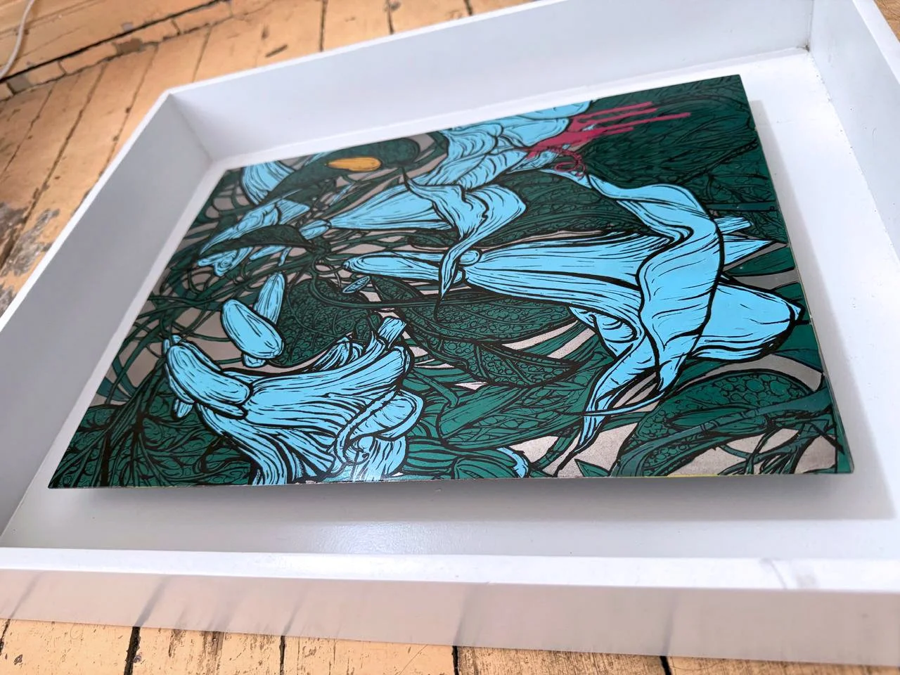 A framed artwork featuring a colorful illustration of flower and plant patterns in shades of blue, green, and pink placed on a wooden surface.