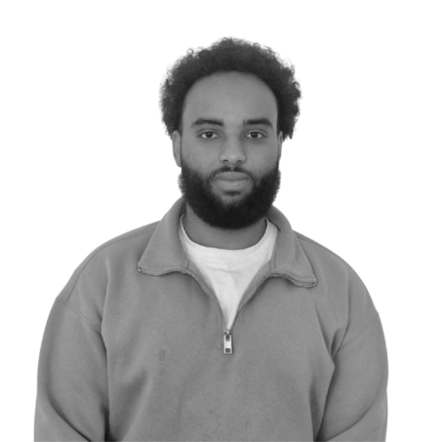 Black man with curly hair and beard, wearing a zip-up sweatshirt and white t-shirt, facing forward against a plain background.