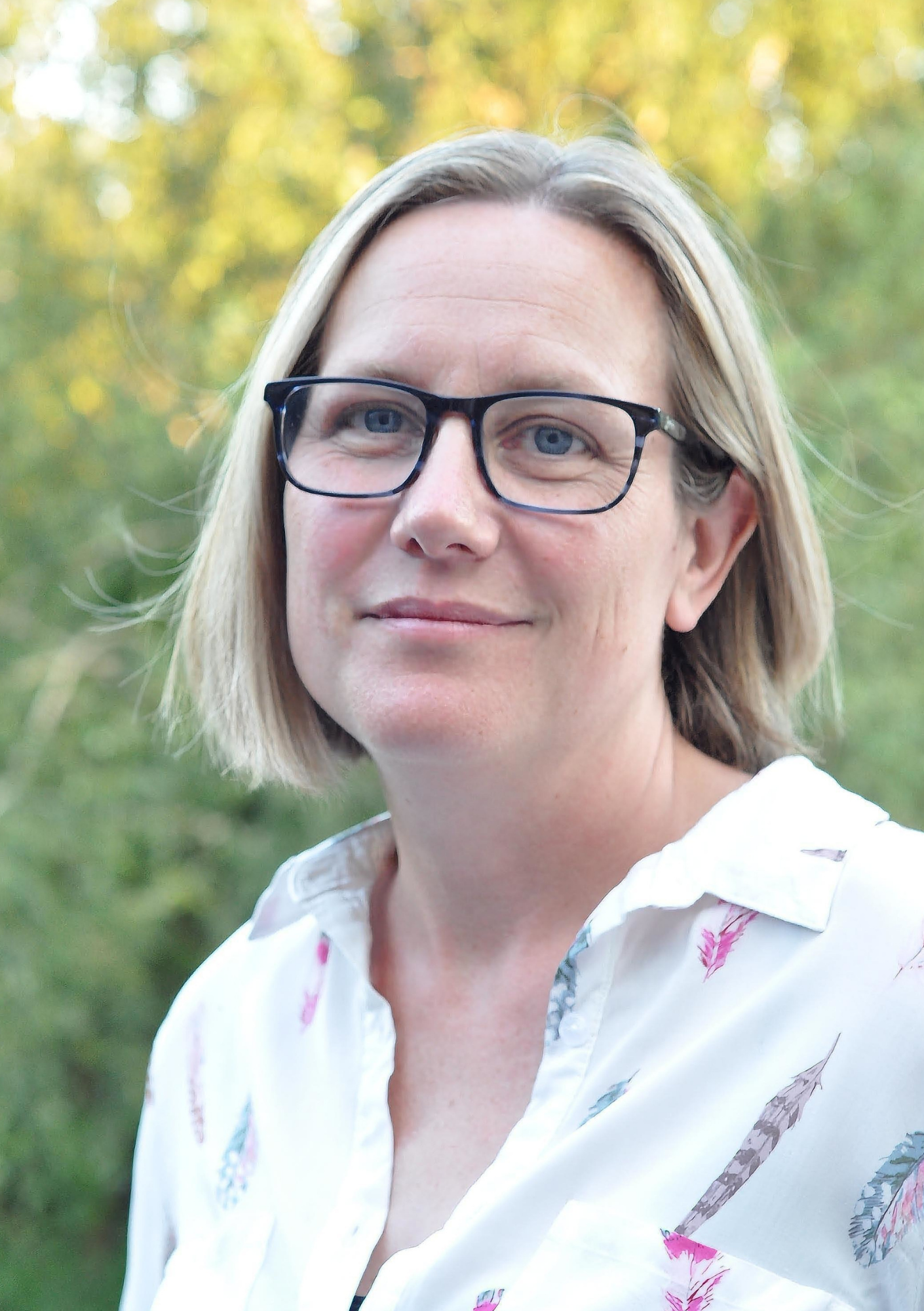 Close-up portrait of a woman with blonde hair, wearing glasses, looking at the camera, outdoors with a blurred background of green foliage.