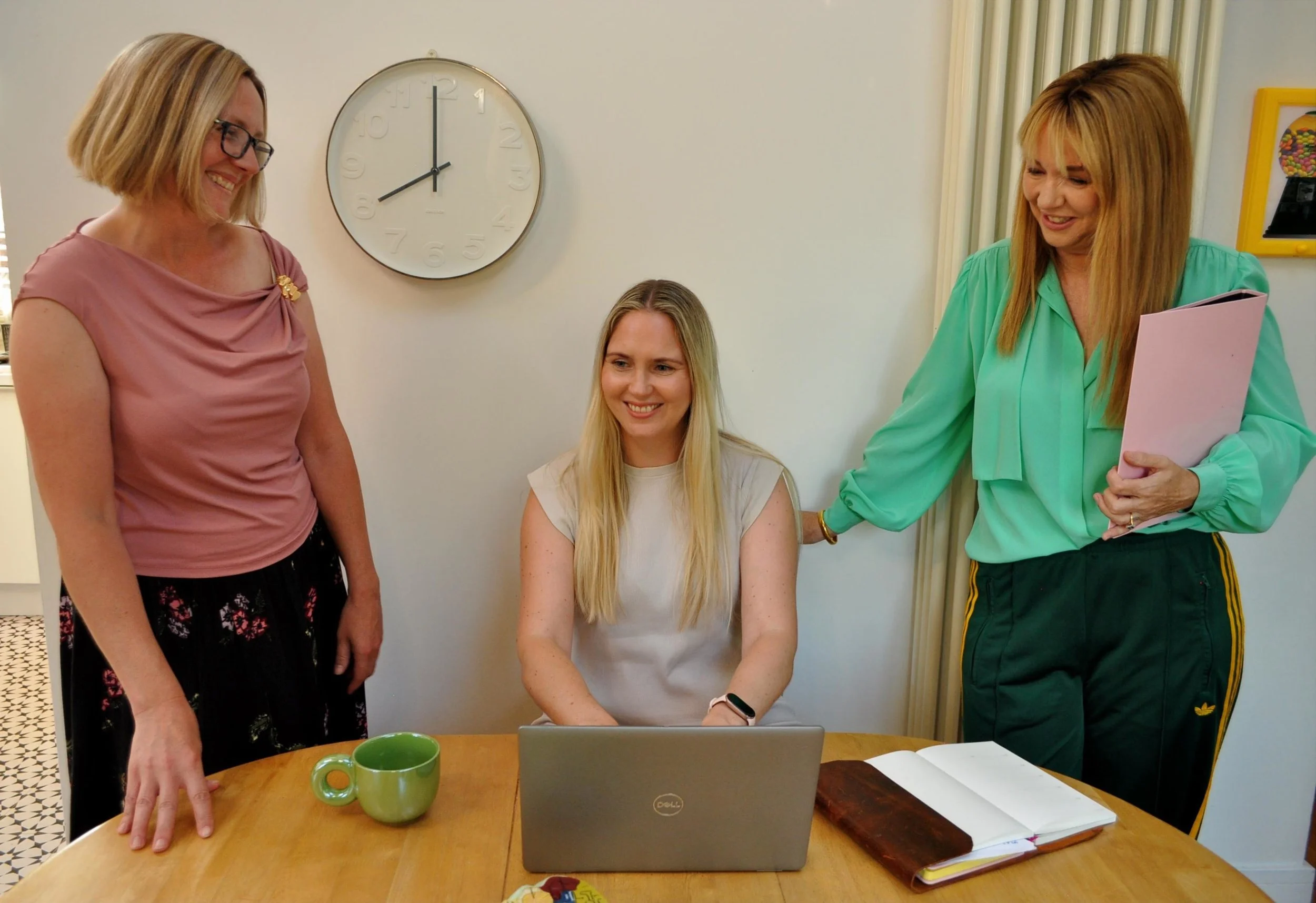 Three women gathered around a laptop at a table in a room with a clock on the wall and a container of colorful candies. The woman in the middle is seated, smiling at the laptop, while the other two women stand beside her, engaged in conversation.