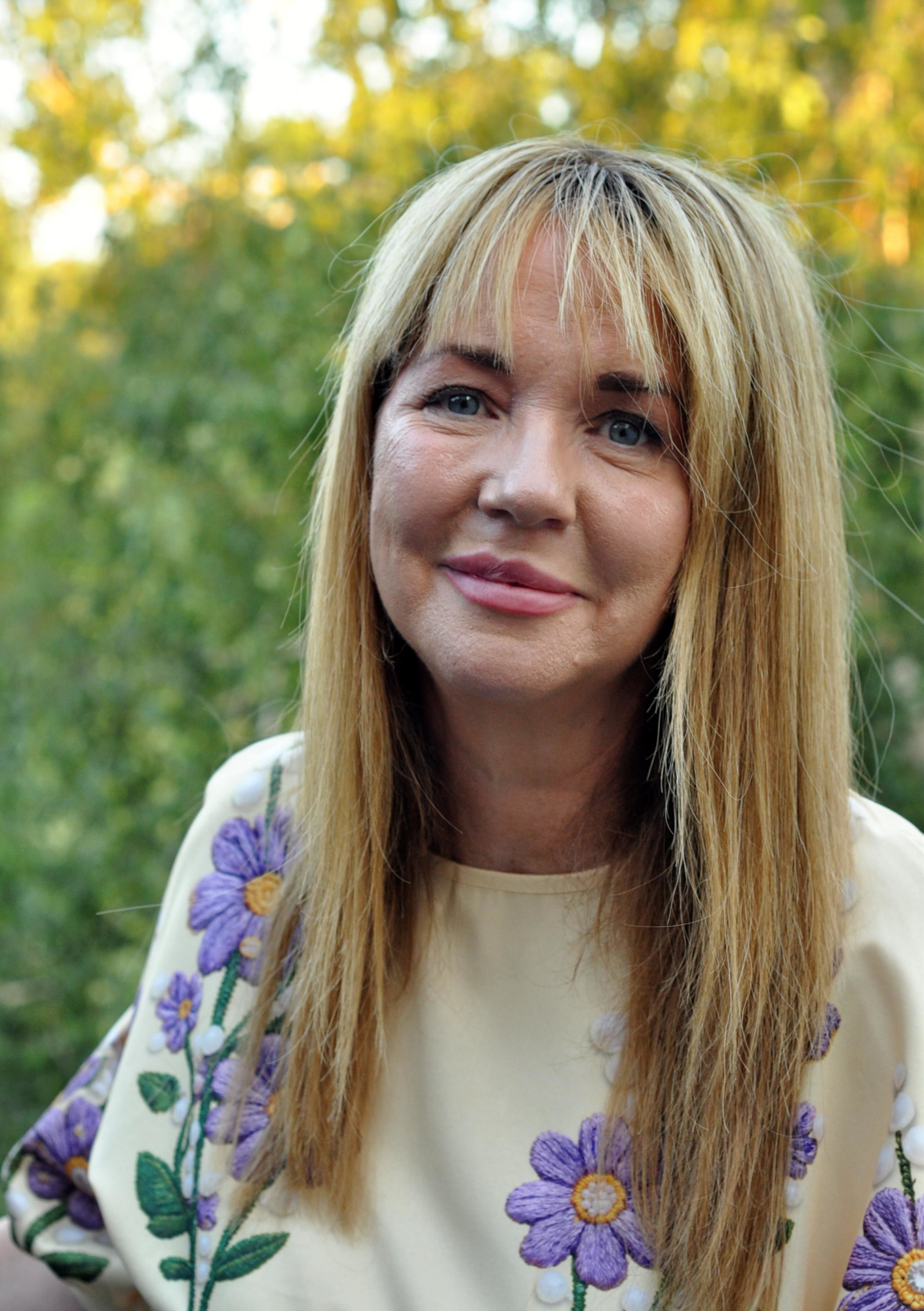 Close-up of a woman with blonde hair and blue eyes, wearing a cream-colored garment with purple embroidered flowers on the sleeves, outdoors with green trees in the background.