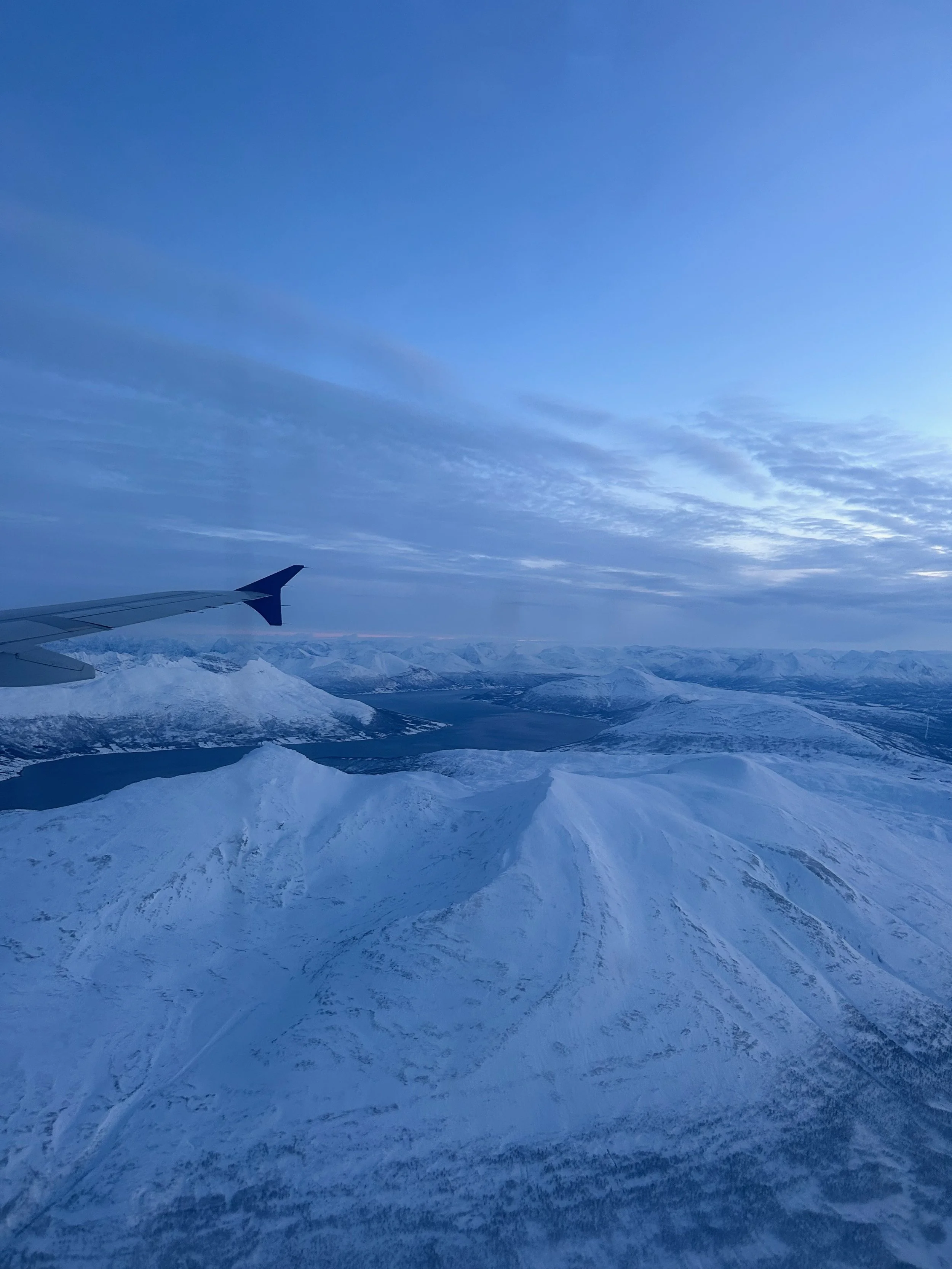Aerial view of snow-covered mountains and lakes seen from an airplane window during daytime with a clear sky and some clouds.