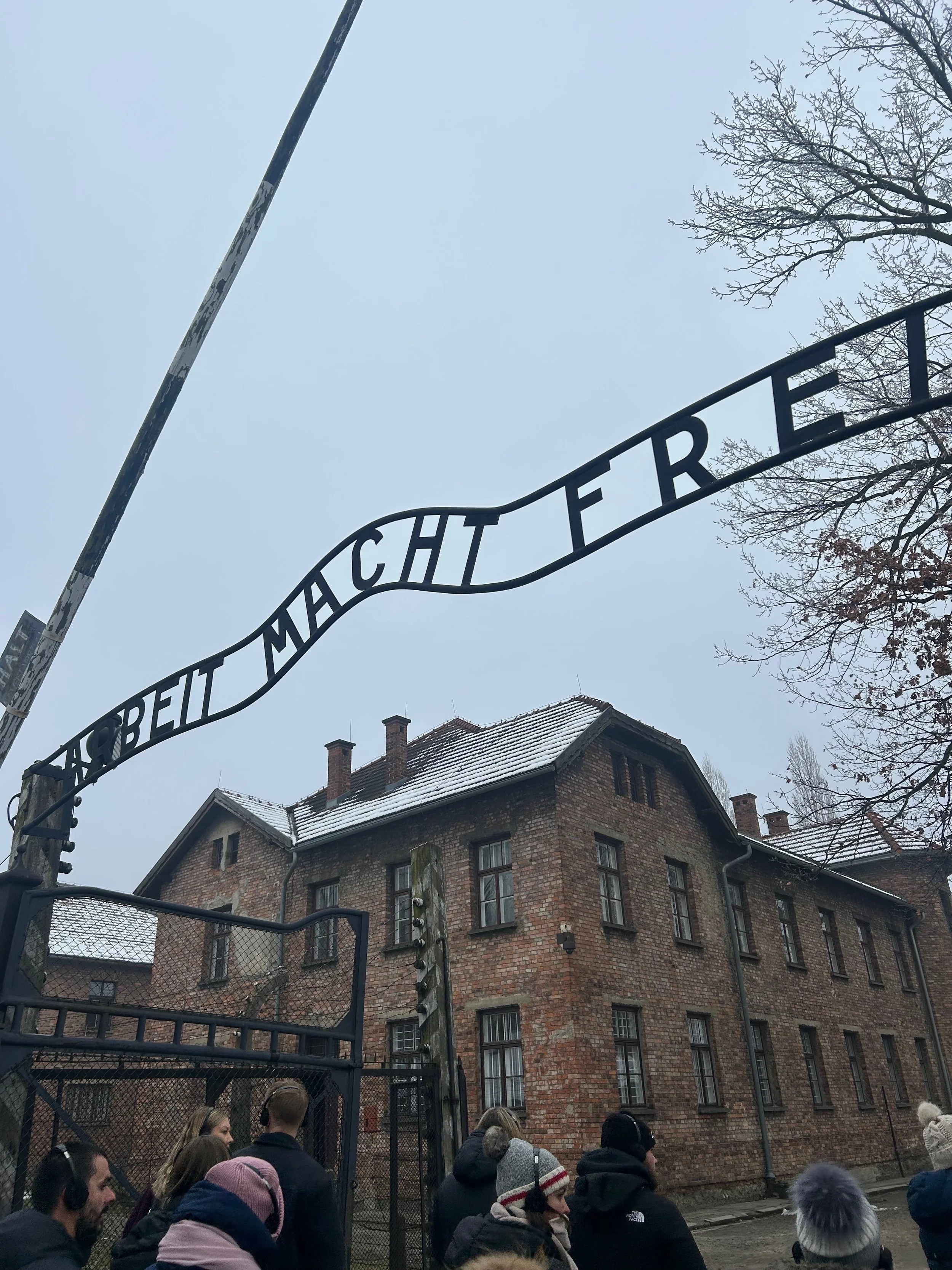 A group of people standing outside the entrance of the Auschwitz concentration camp, with the iconic 'Arbeit Macht Frei' sign arching over the gate and a brick building in the background.