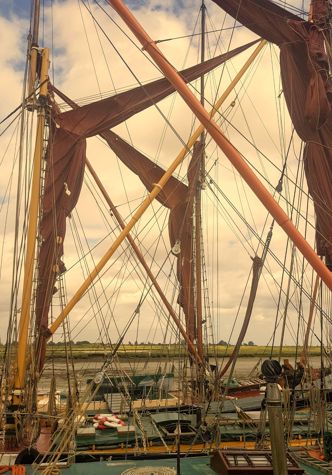 View of a tall ship's masts, sails, and rigging during daytime with a partly cloudy sky and green landscape in the background.
