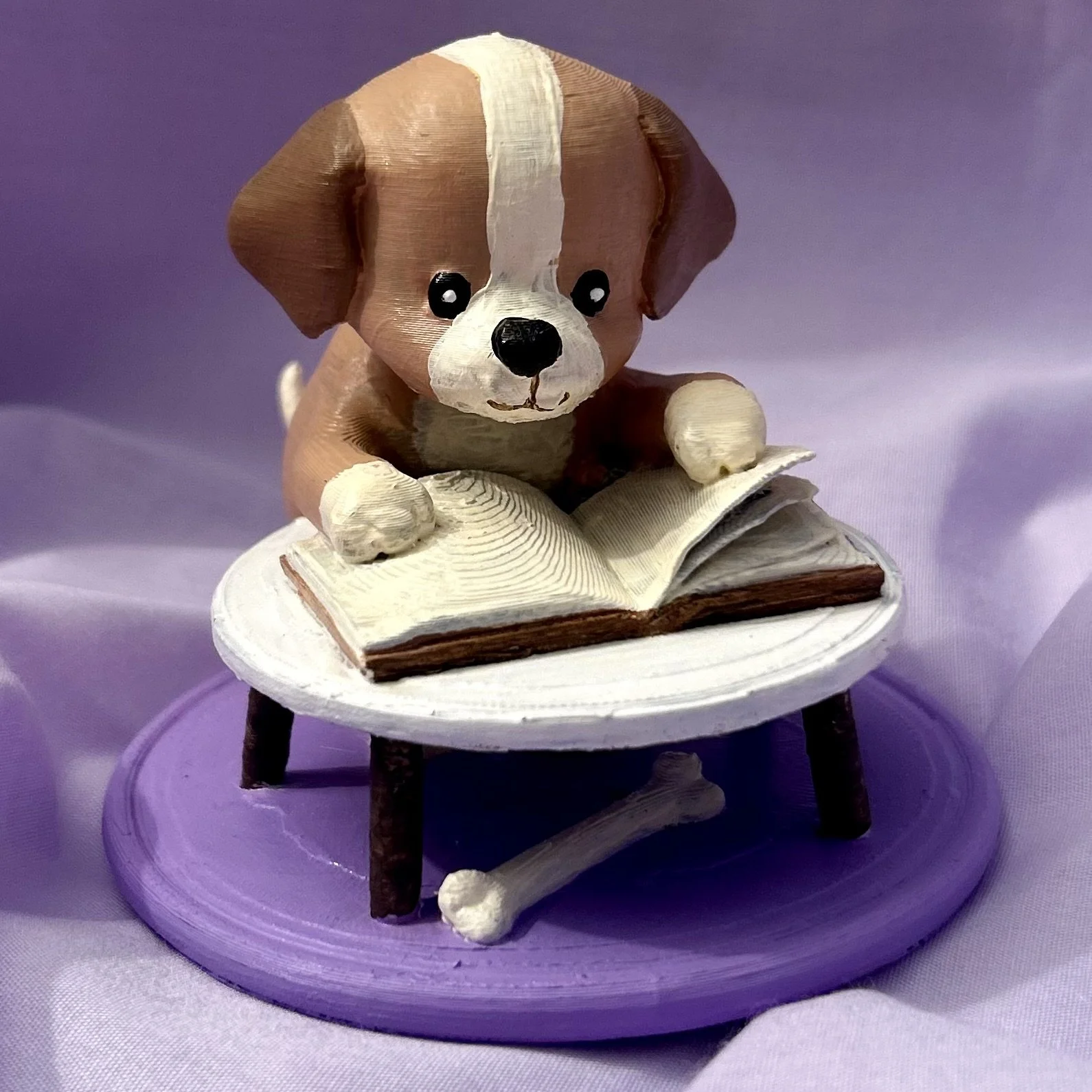 Handcrafted 3D figurine of Toby the Puppy Page Turner sitting at a small table reading a book, displayed on a soft lilac fabric background.