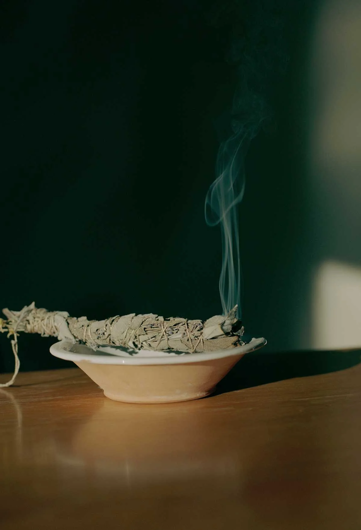 A smoldering bundle of sage on a dish, with smoke rising in a gentle swirl, on a wooden surface against a dark background.
