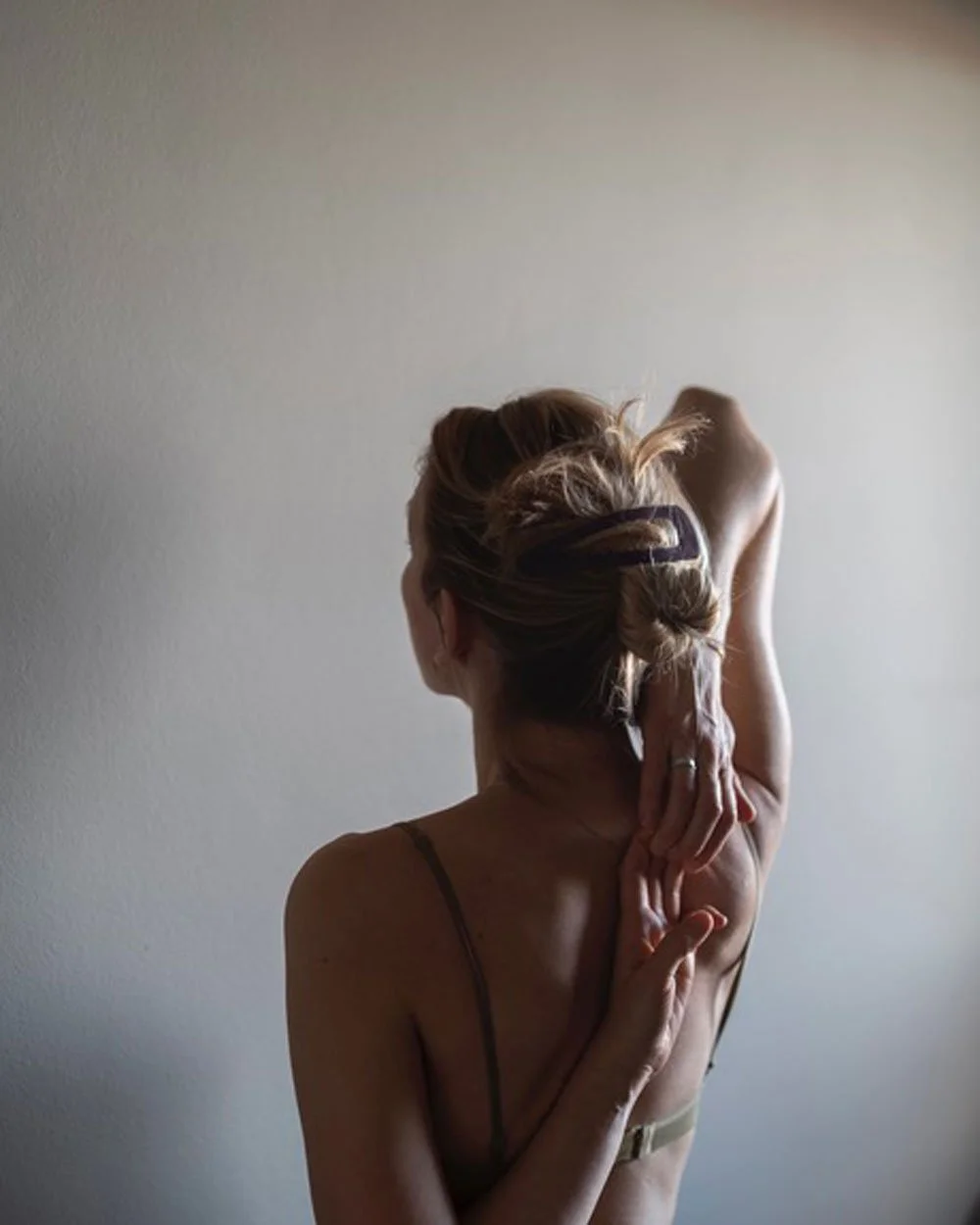 A woman with brown hair in a bun stretching her arm behind her head, standing against a plain wall.
