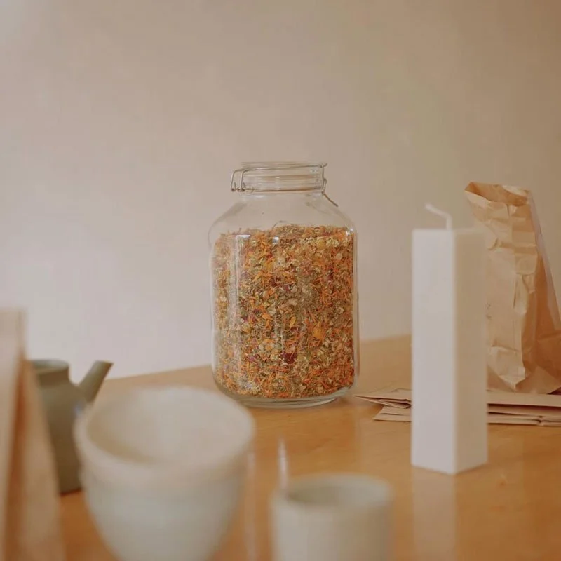 A glass jar filled with dried herbal or spice mixture on a wooden table, with white candles, a teapot, paper bag, and papers around it.