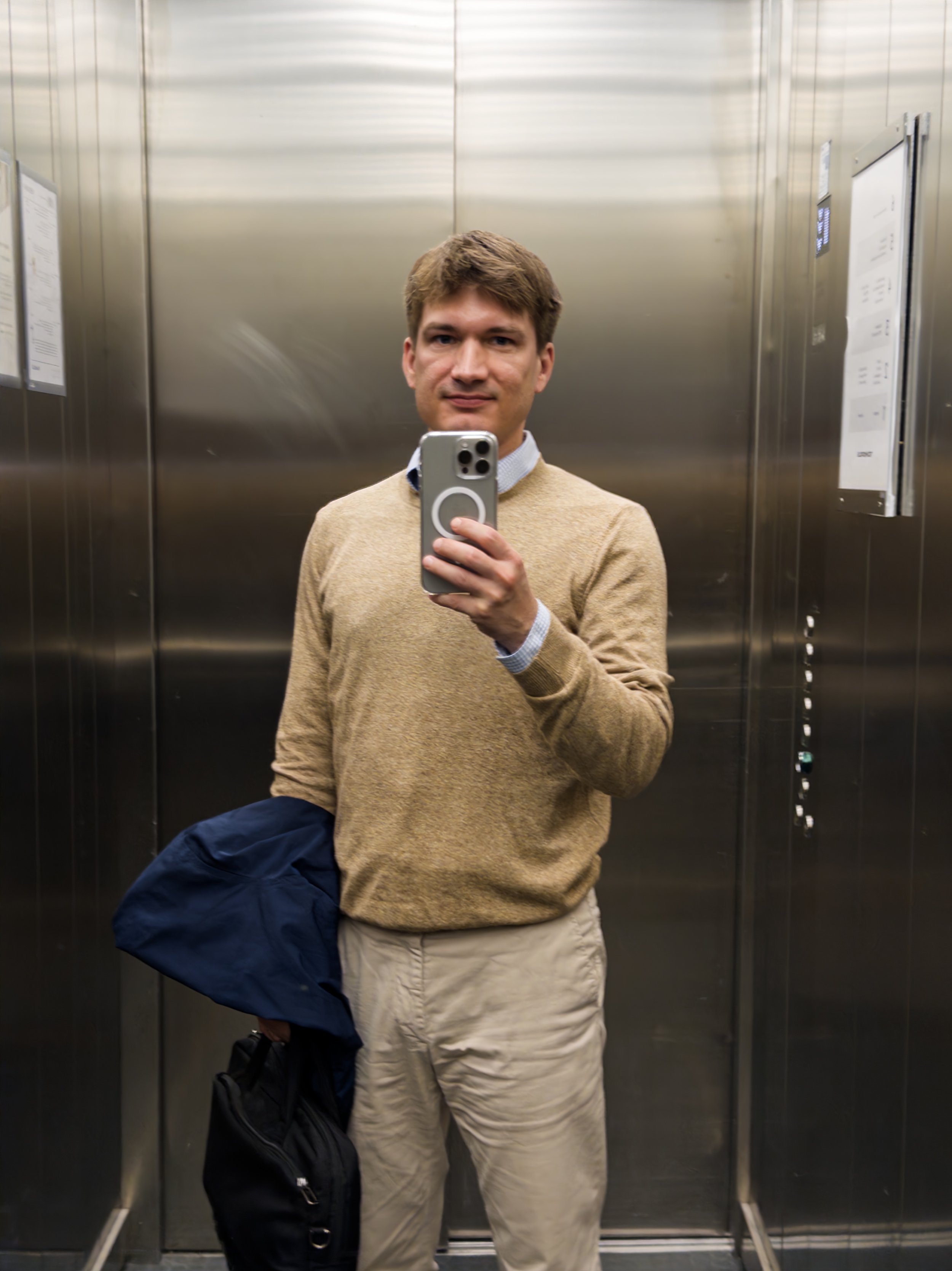 Nuorehko mies ottaa selfietä hissin peilistä, hänellä on ruskea lyhyt tukka, beige pusero ja vaaleat housut, hänellä on käsissään pusero ja reppu.