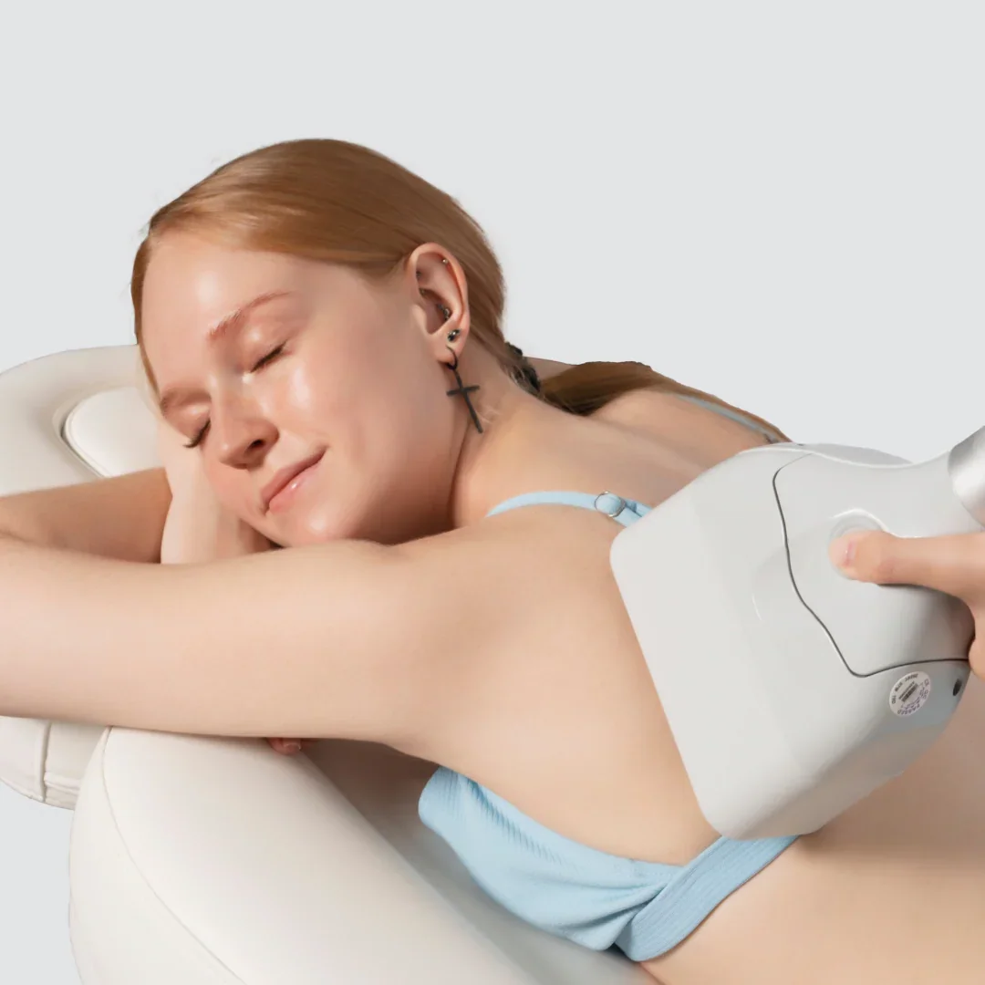 A woman with red hair and multiple earrings, including a cross earring, is receiving a cosmetic or medical treatment on her upper arm with a device, while she relaxes with her eyes closed.