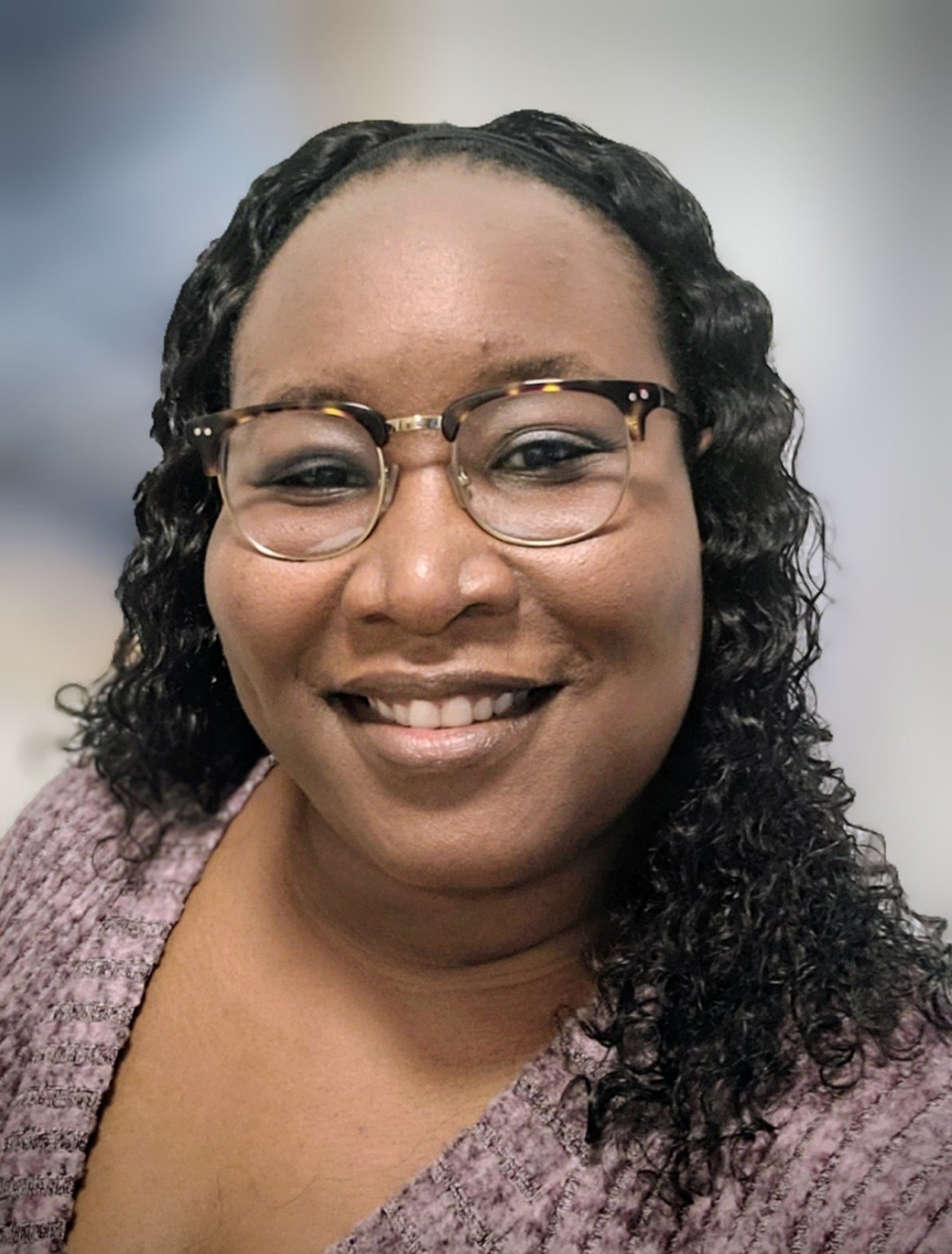 Close-up of a woman with glasses, curly hair, and a warm smile, wearing a purple textured top, against a neutral background.