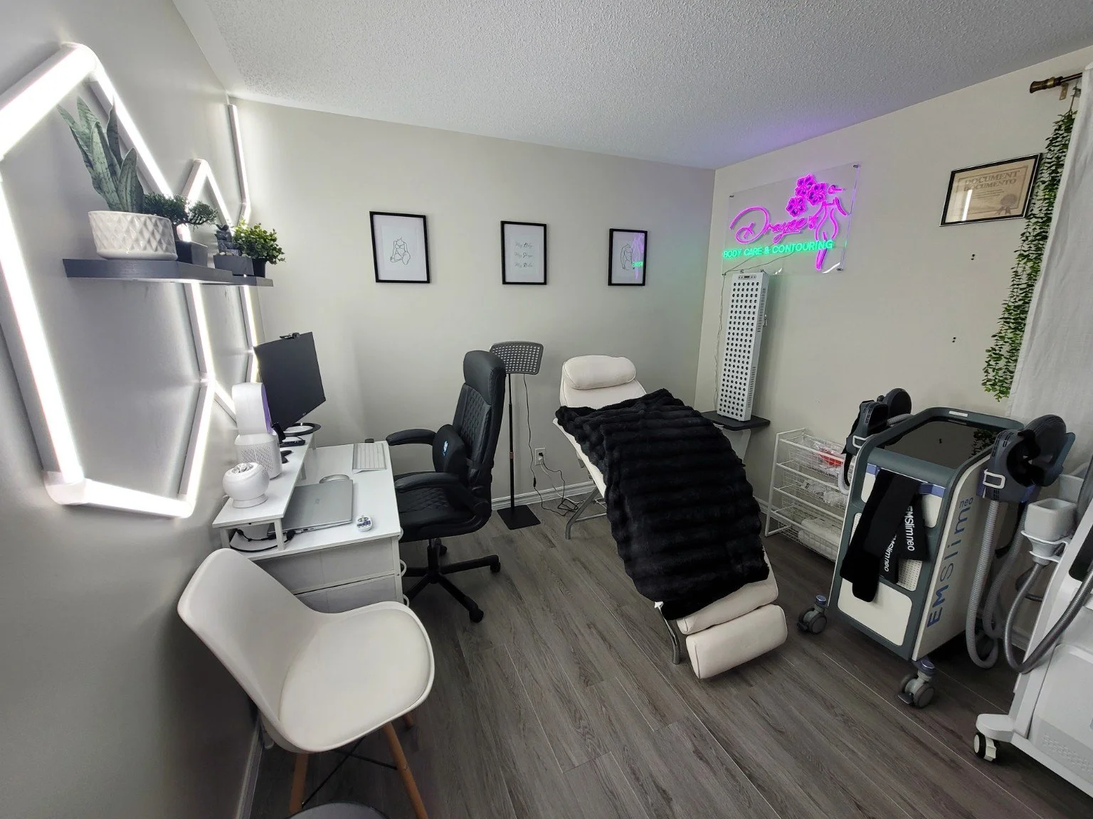A room with a treatment bed covered with a black blanket, a computer desk with a monitor and chair, and various medical or beauty equipment. The wall has framed pictures and a neon sign that says 'Dragon' and 'Body Care & Contouring.' There are shelv