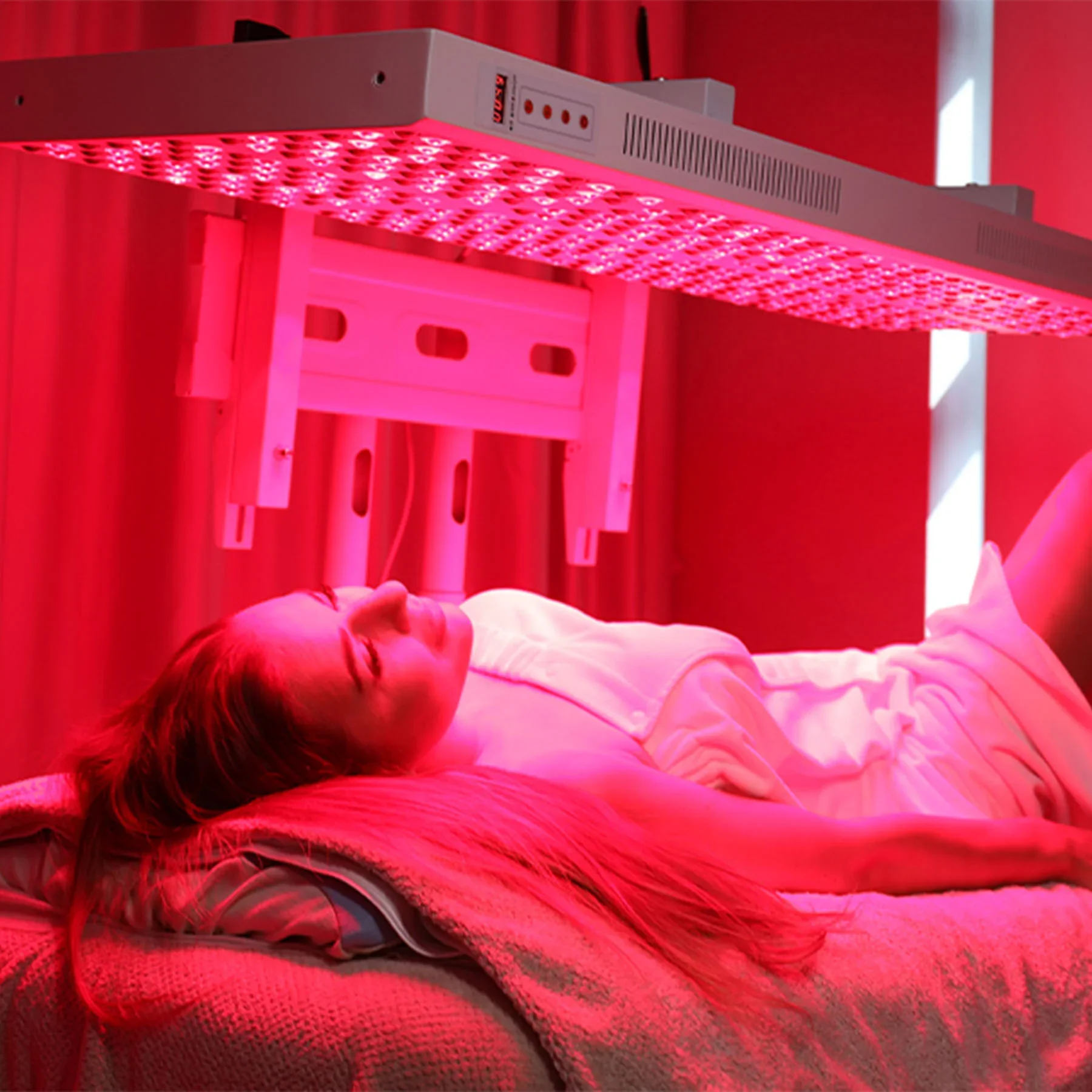 A woman lying on a bed under a pink grow light in a red-lit room.