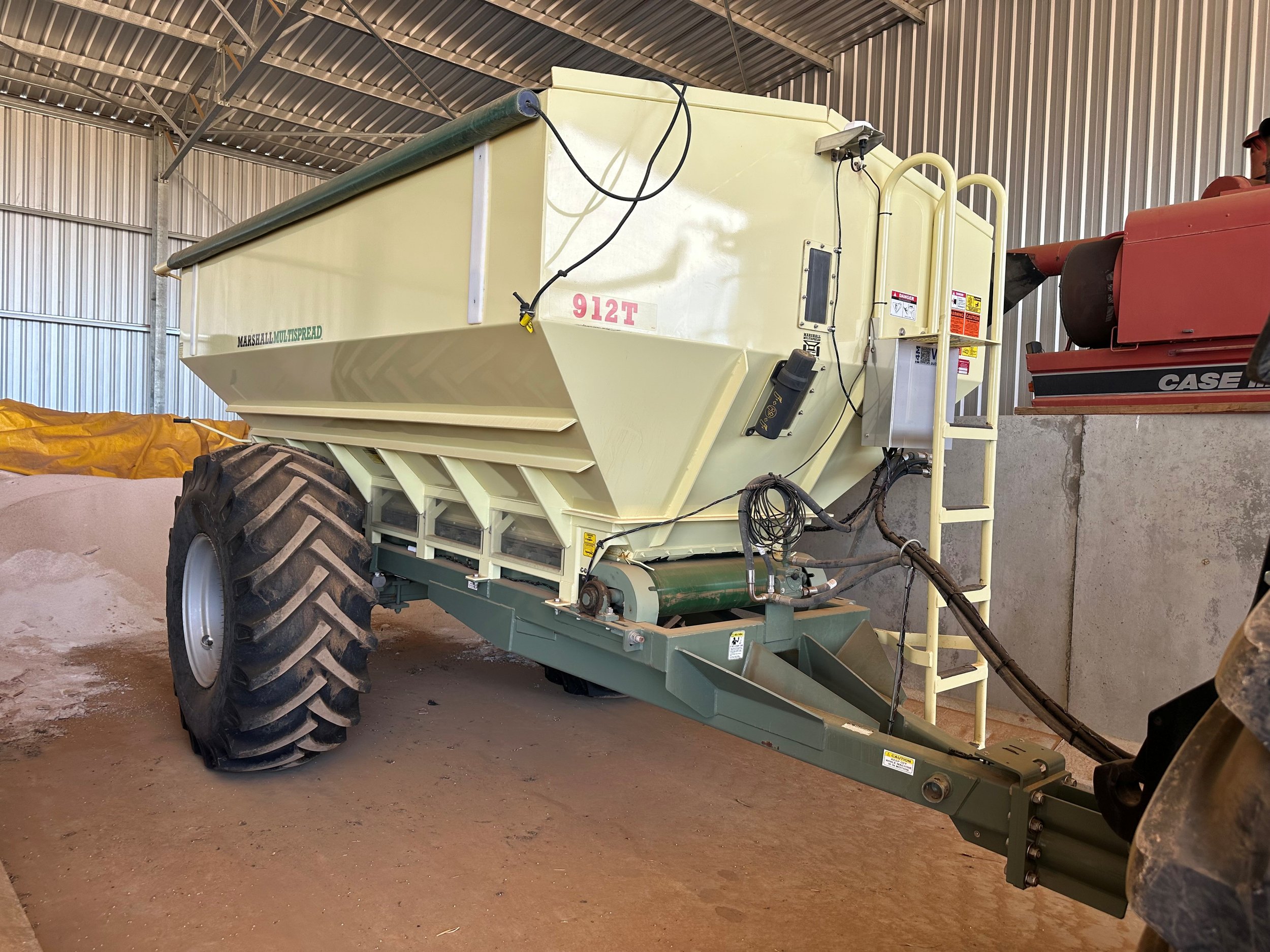 A large agricultural spreader or fertilizer applicator inside a barn, with large black tires, connected via hydraulic hoses to a tractor.