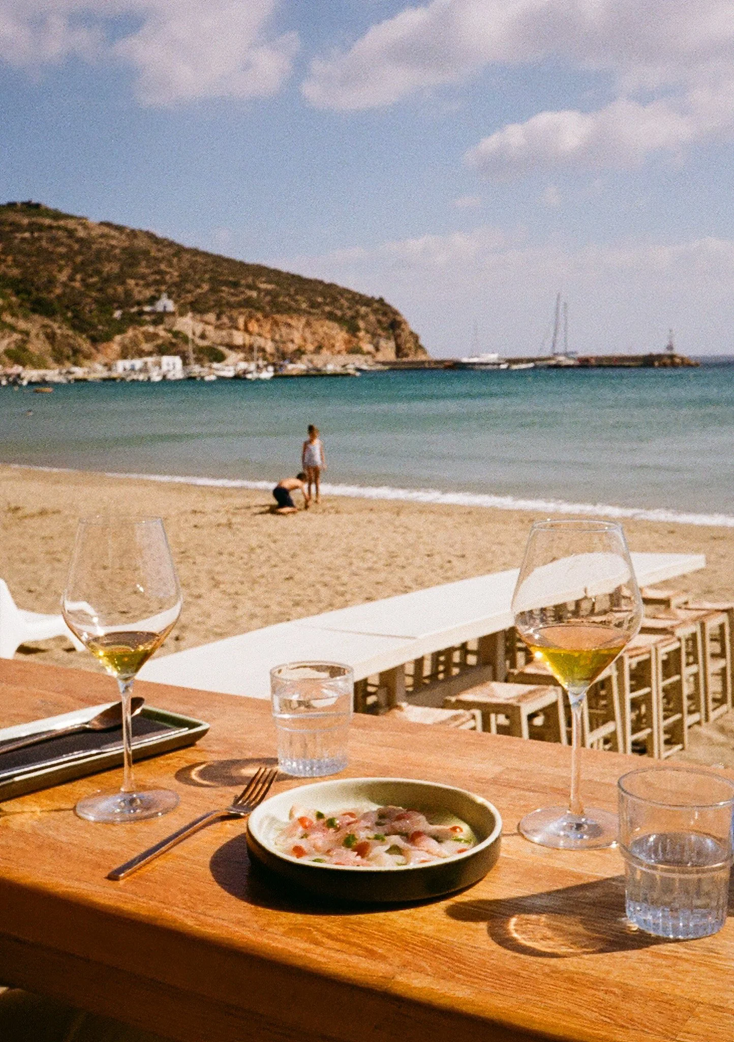 A beachside dining scene with a table set with wine glasses, a bowl of food, and water glasses, overlooking a sandy beach with a boat, a large hill, and a cloudy sky in the background.