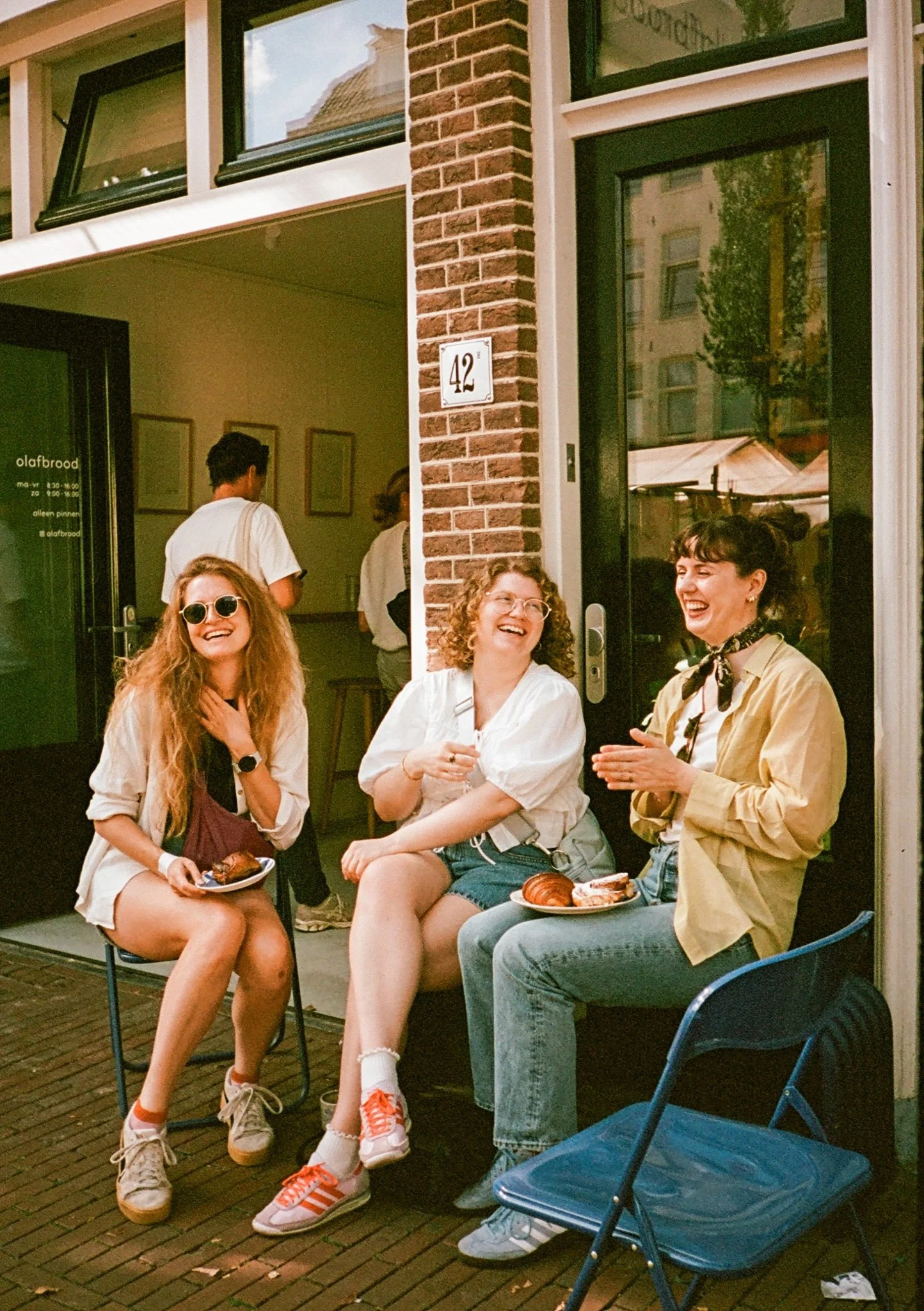 Three women sitting outside a cafe, laughing and enjoying pastries, with an open doorway behind them showing two more people inside.