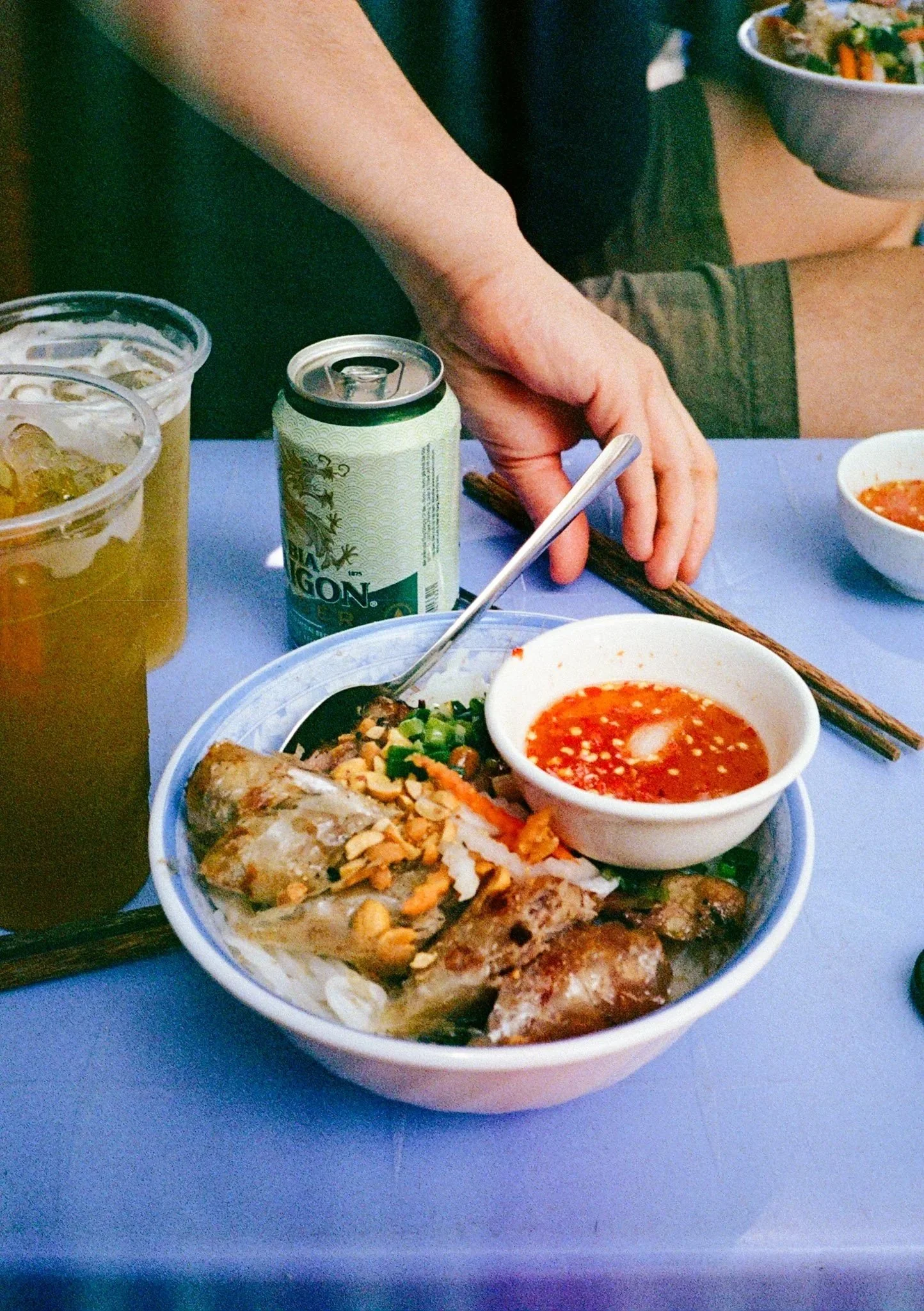 Bowl of Vietnamese food with spring rolls, and a small bowl of red chili sauce, on a table with drinks and chopsticks.