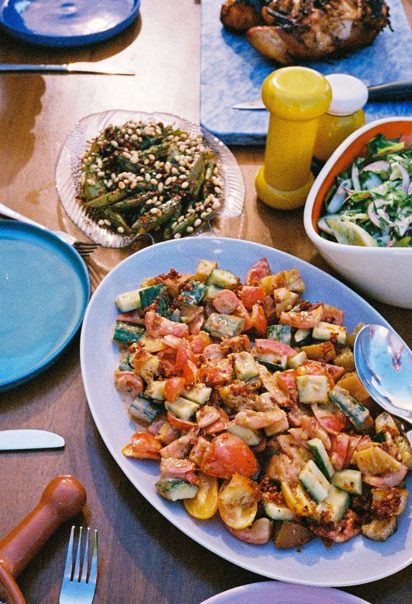 A table with various dishes including a large plate of tomato and cucumber salad, a bowl of green bean salad with pine nuts, a mixed vegetable dish, and a bowl of salad with greens and onion slices, with condiments and utensils.