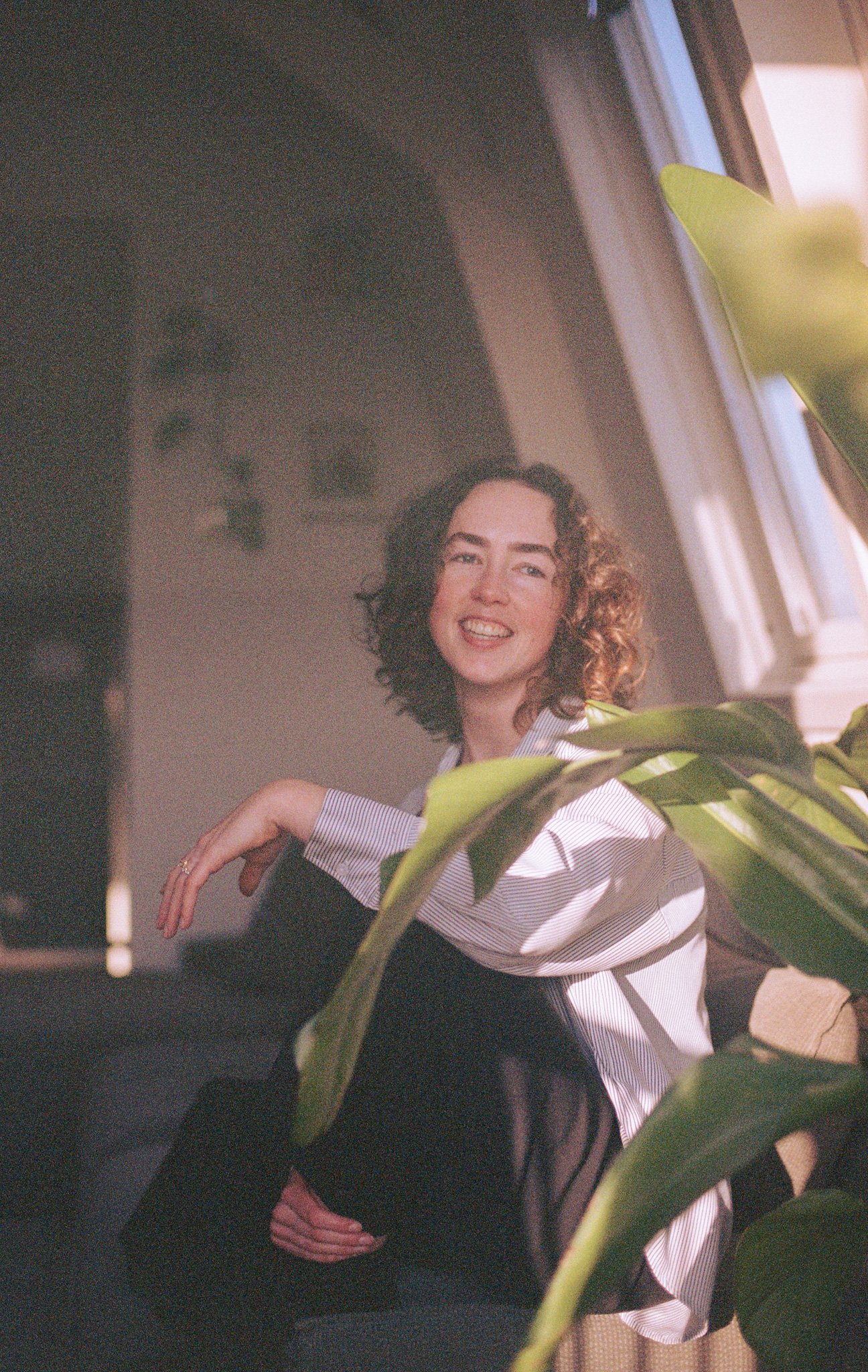 A woman with curly hair smiling while sitting near a window with sunlight, partially obscured by large green plant leaves.