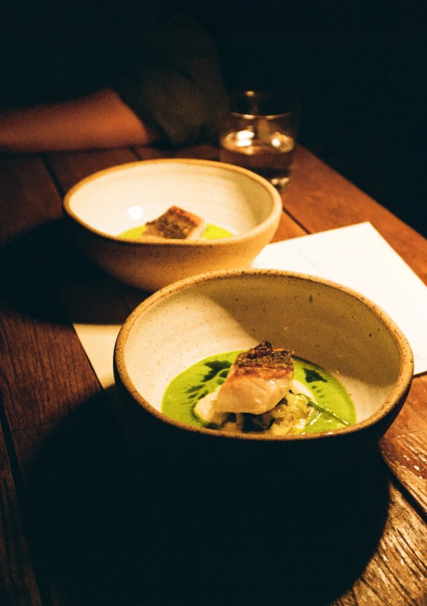 Two bowls of gourmet food on a wooden table, each containing a small piece of grilled meat on a bed of green sauce or puree.