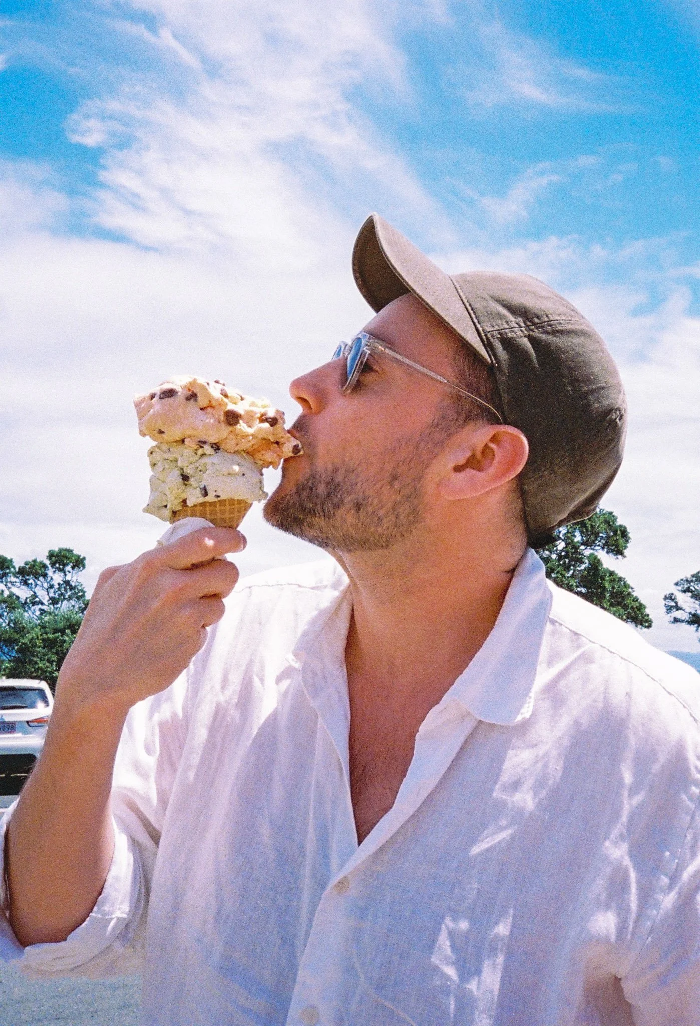 A man wearing sunglasses, a cap, and a white shirt eating a large ice cream cone outdoors against a blue sky with clouds.
